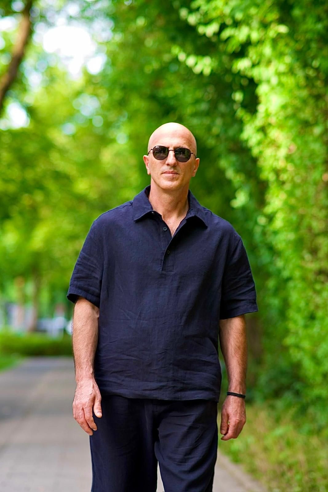 Bald man in sunglasses wearing a navy blue linen outfit walking on a tree-lined green pathway.