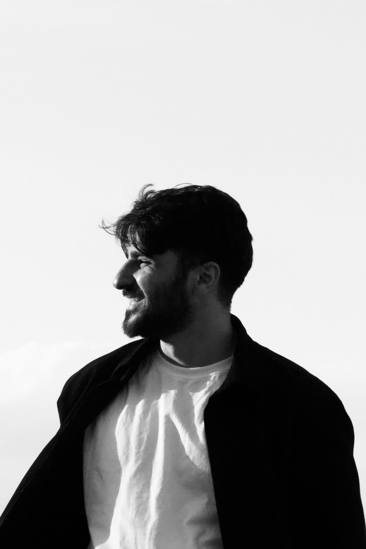 Black and white portrait of a smiling bearded man in a white t-shirt and dark jacket outdoors.