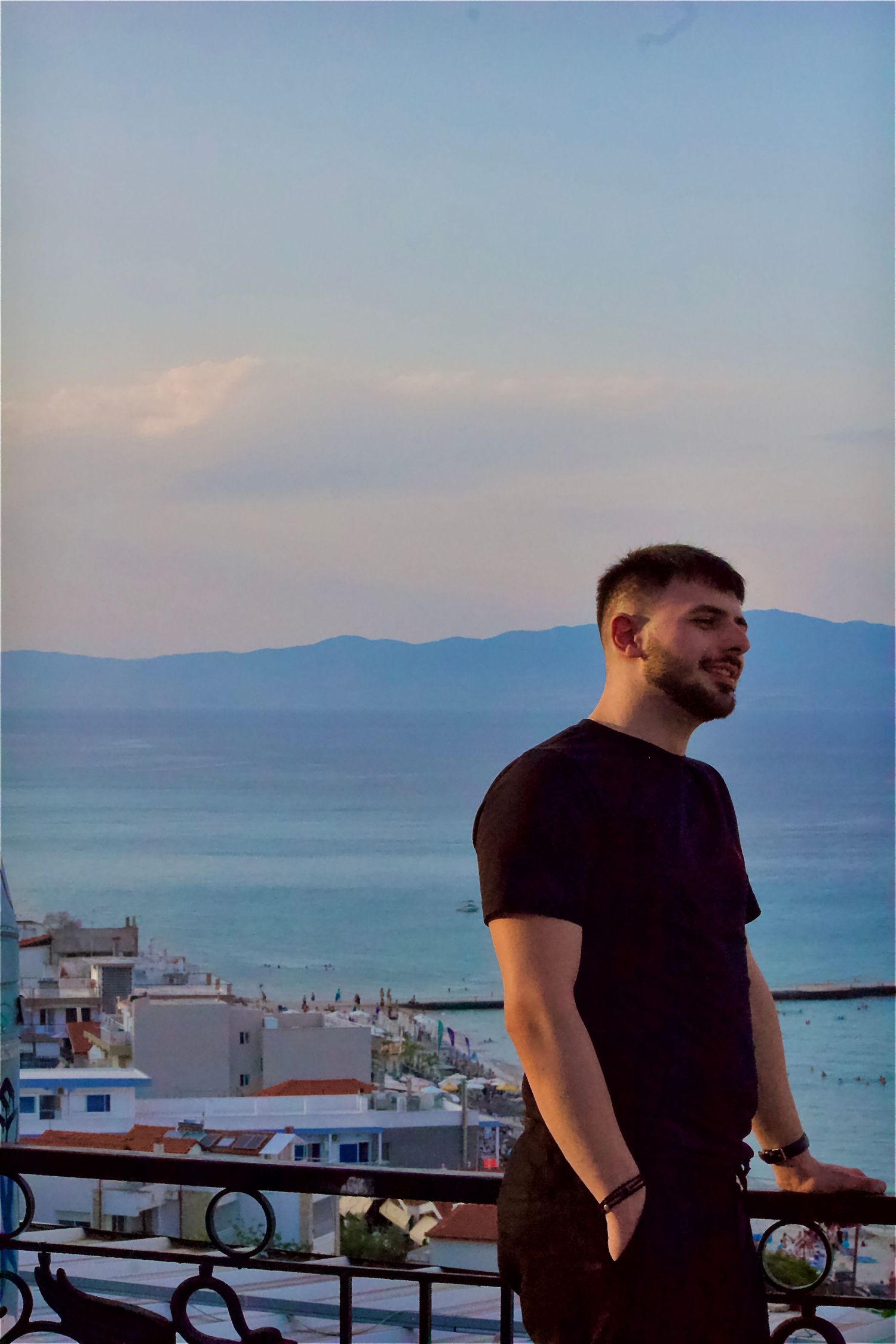 Young man in black t-shirt smiling on a balcony with scenic ocean and mountain views at sunset.