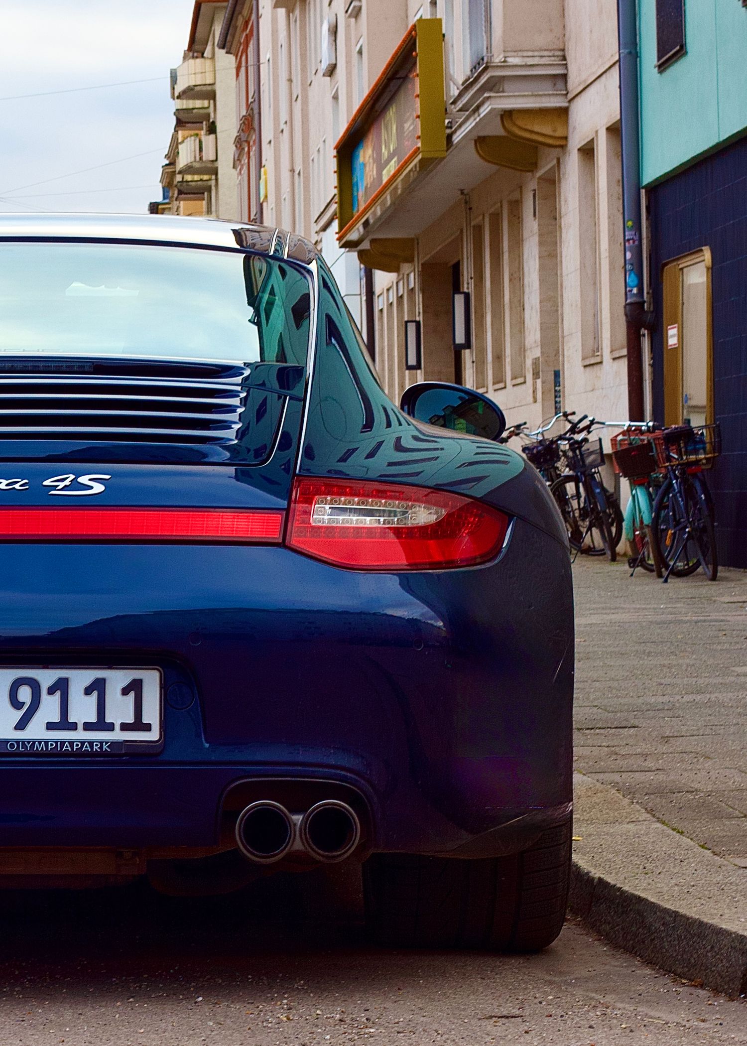 Blue Porsche 911 4S sports car parked on a city street with buildings and bicycles in background.