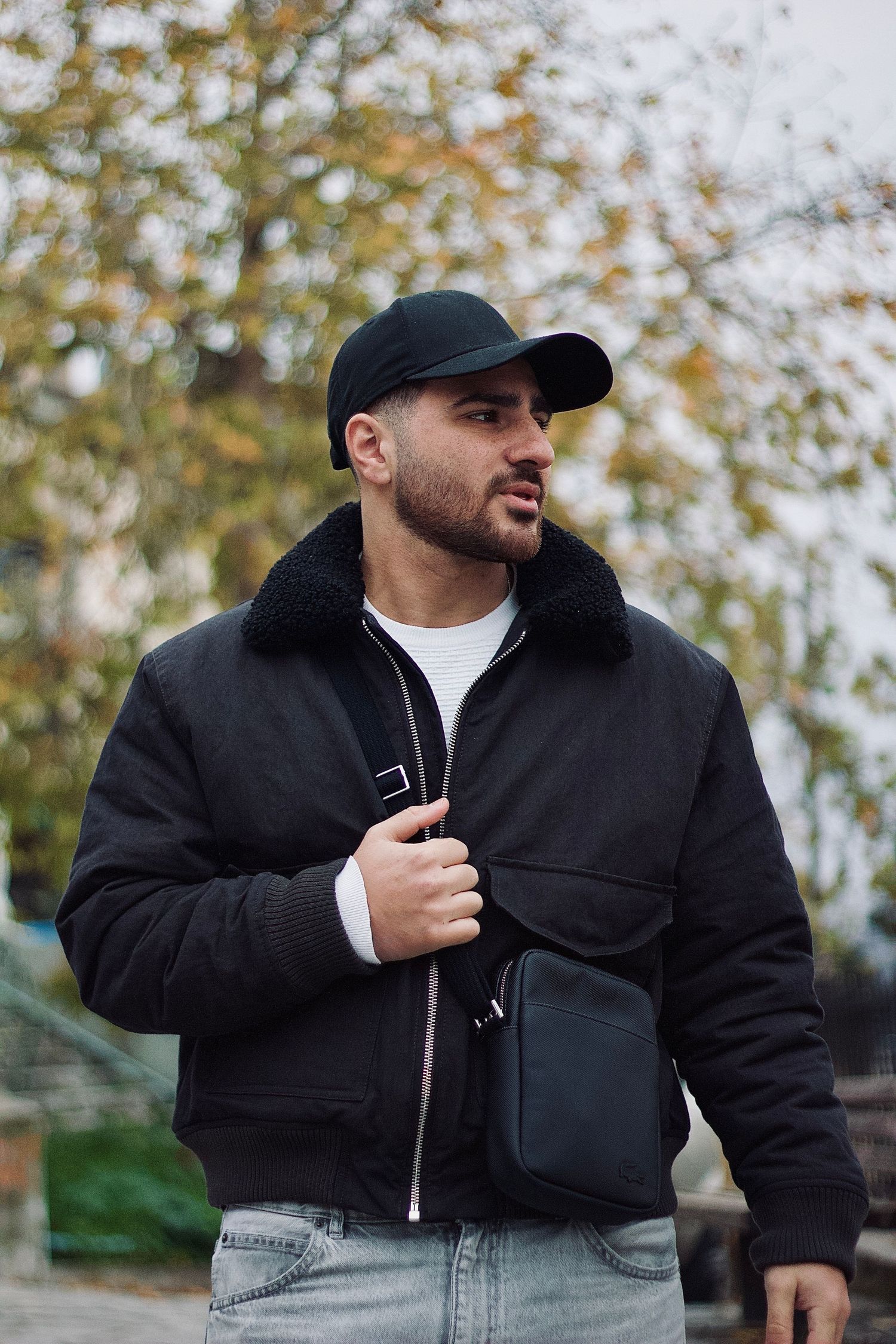 Bearded man in black bomber jacket and cap holds crossbody bag outdoors among autumn trees.