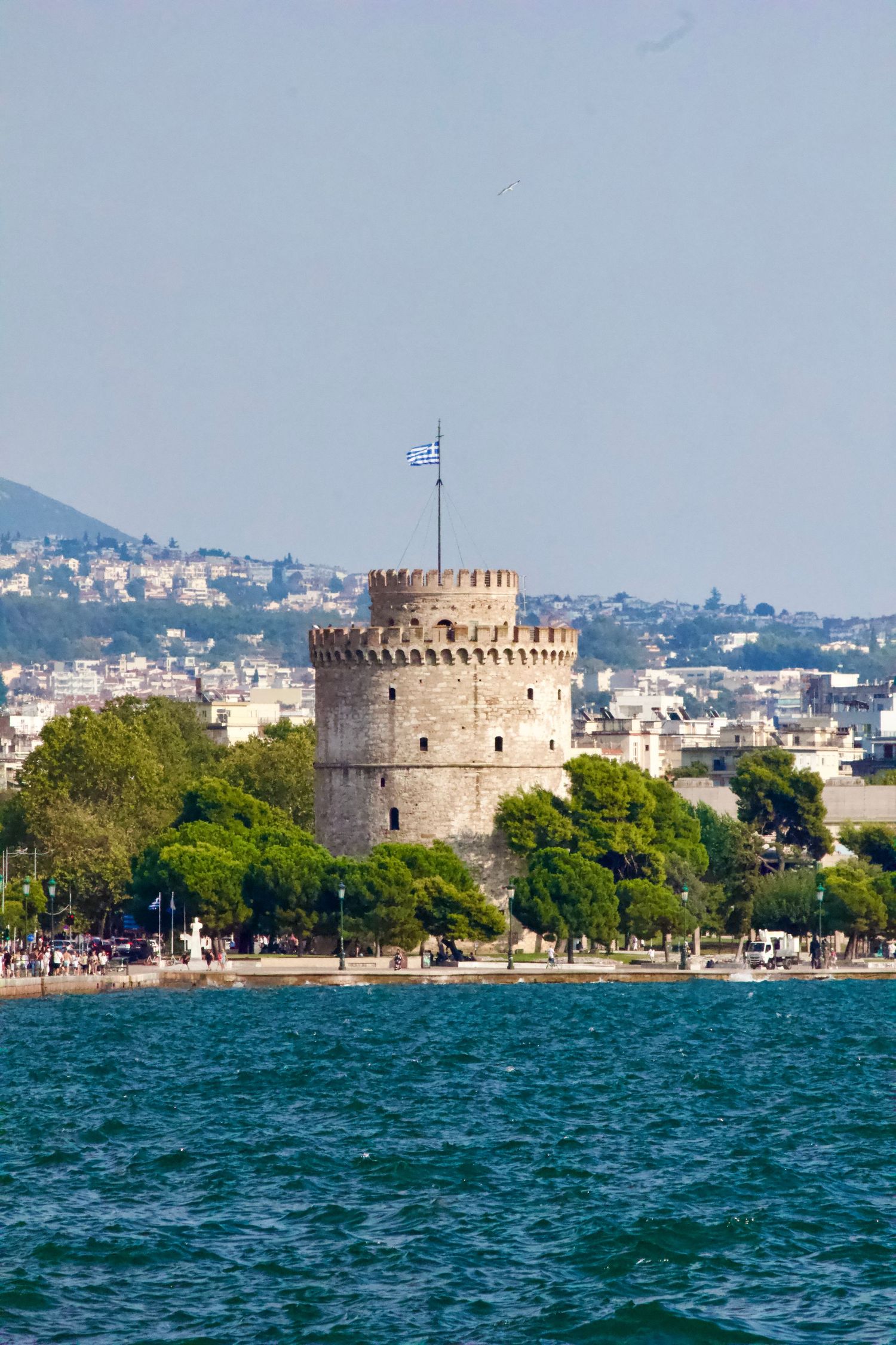 The White Tower of Thessaloniki with Greek flag beside the blue sea with city in background.