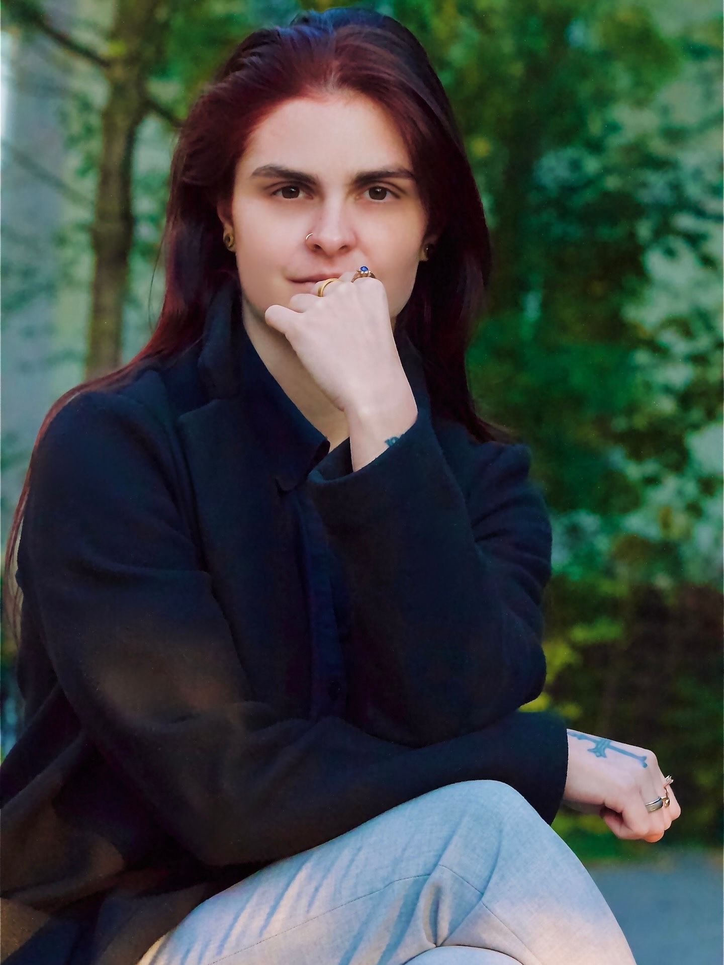 Young woman with long red hair wearing a black blazer sits outdoors with hand near chin in thoughtful pose.