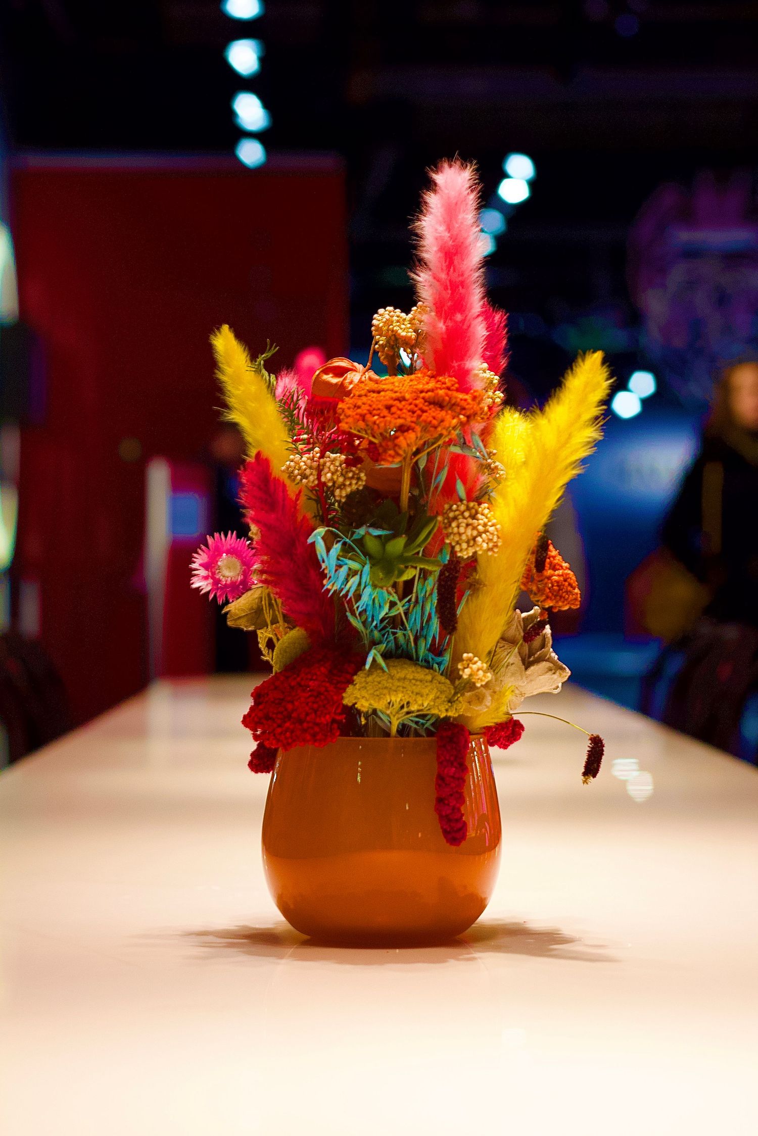Vibrant colorful dried flower arrangement in a brown vase on a white table with bokeh lights in background.