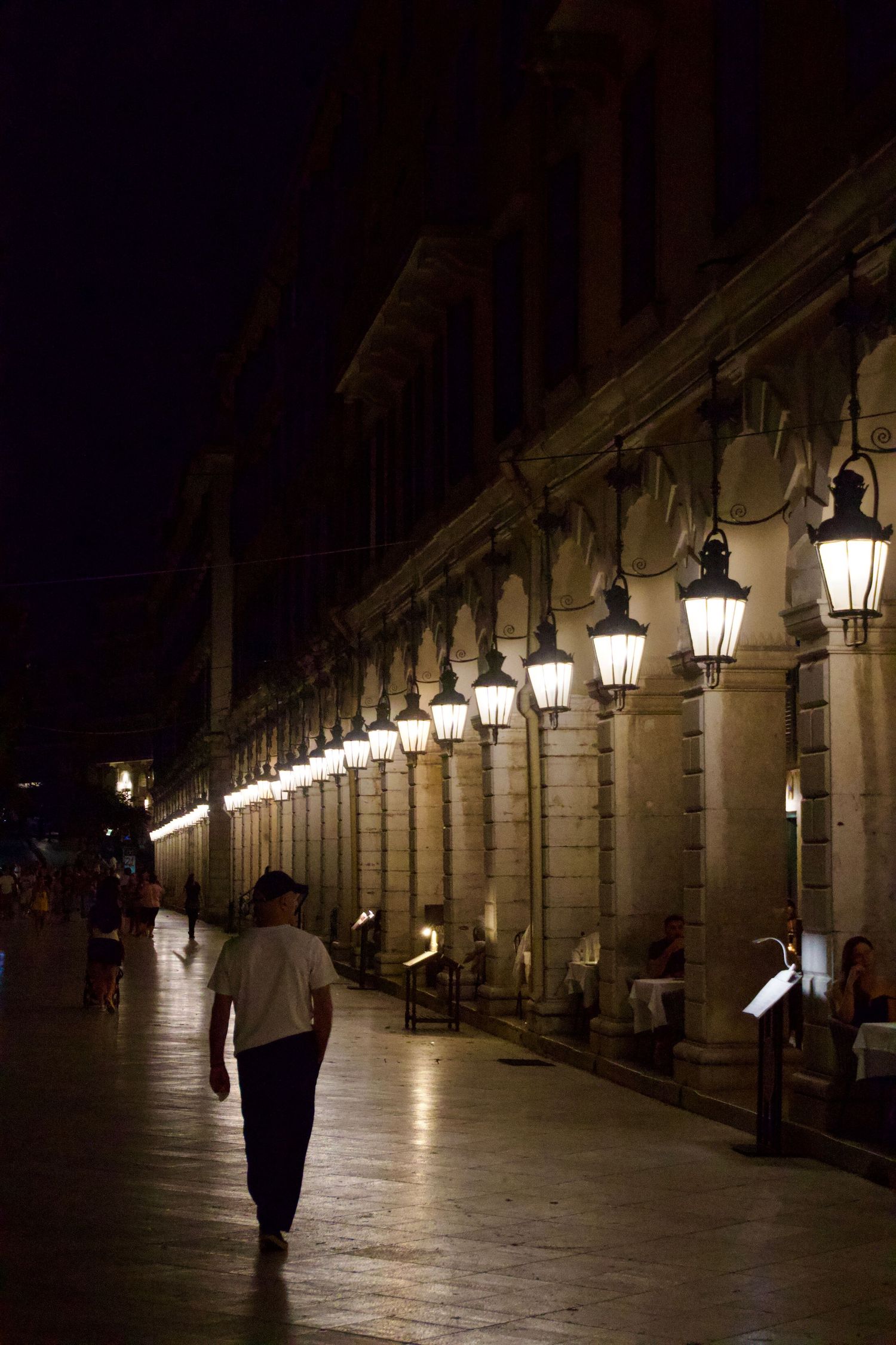 Man walking along illuminated colonnade with glowing lanterns on historic street at night.