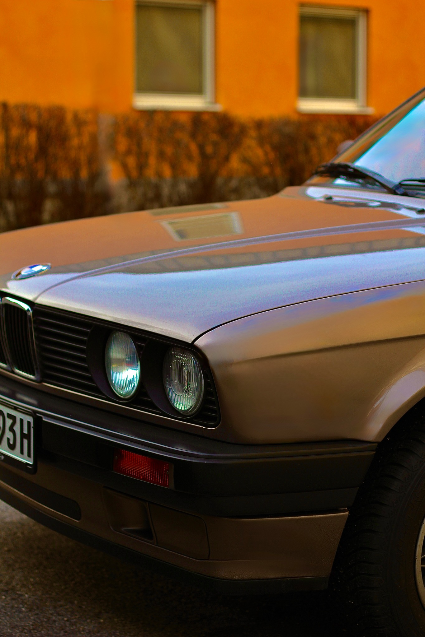 Front corner of a vintage BMW E30 in bronze color parked against an orange building wall.