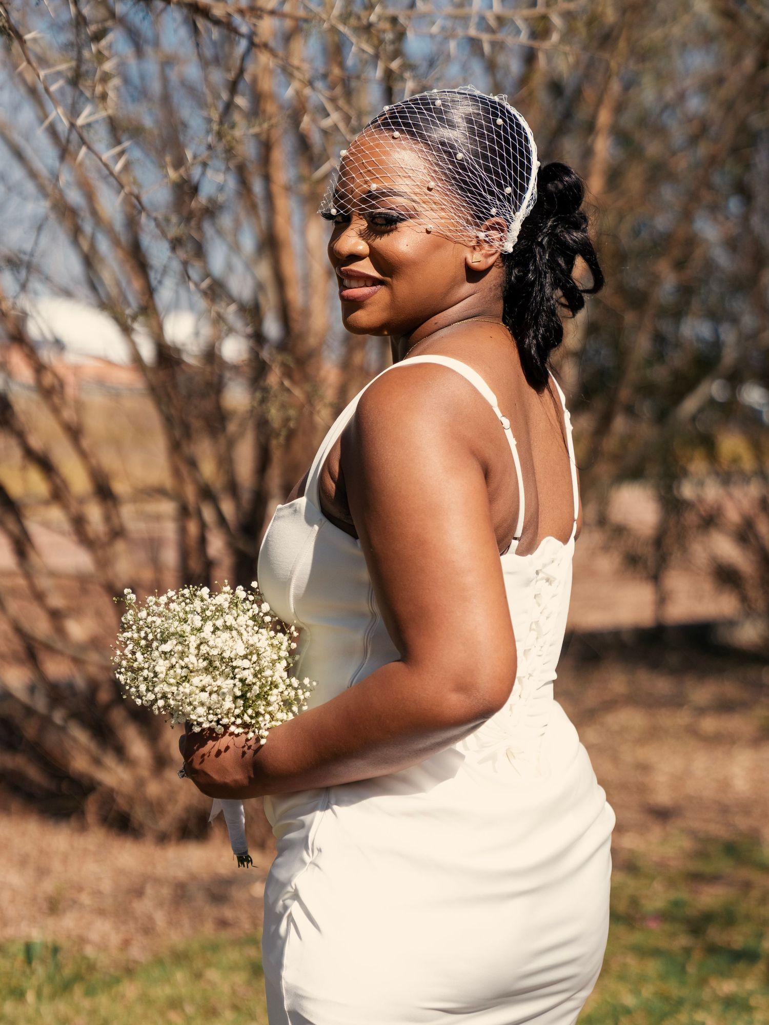 A smiling bride in a white gown holds a baby's breath bouquet, wearing a birdcage veil outdoors. Wedding Photography