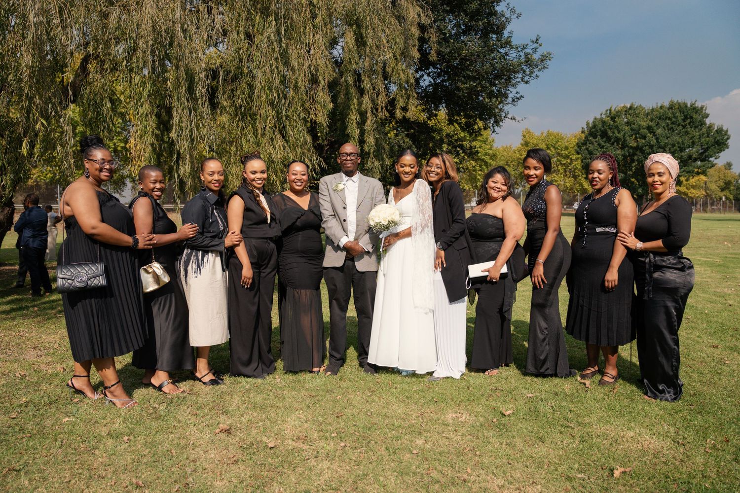 Wedding couple poses with group of guests dressed in black attire under willow tree. Wedding Photography