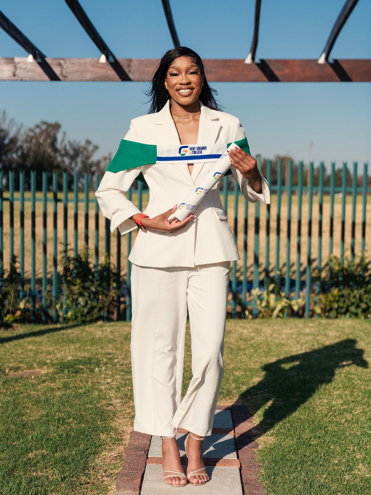 Confident graduate in white suit holding diploma in sunny outdoor setting. Babyshower Photoshoot