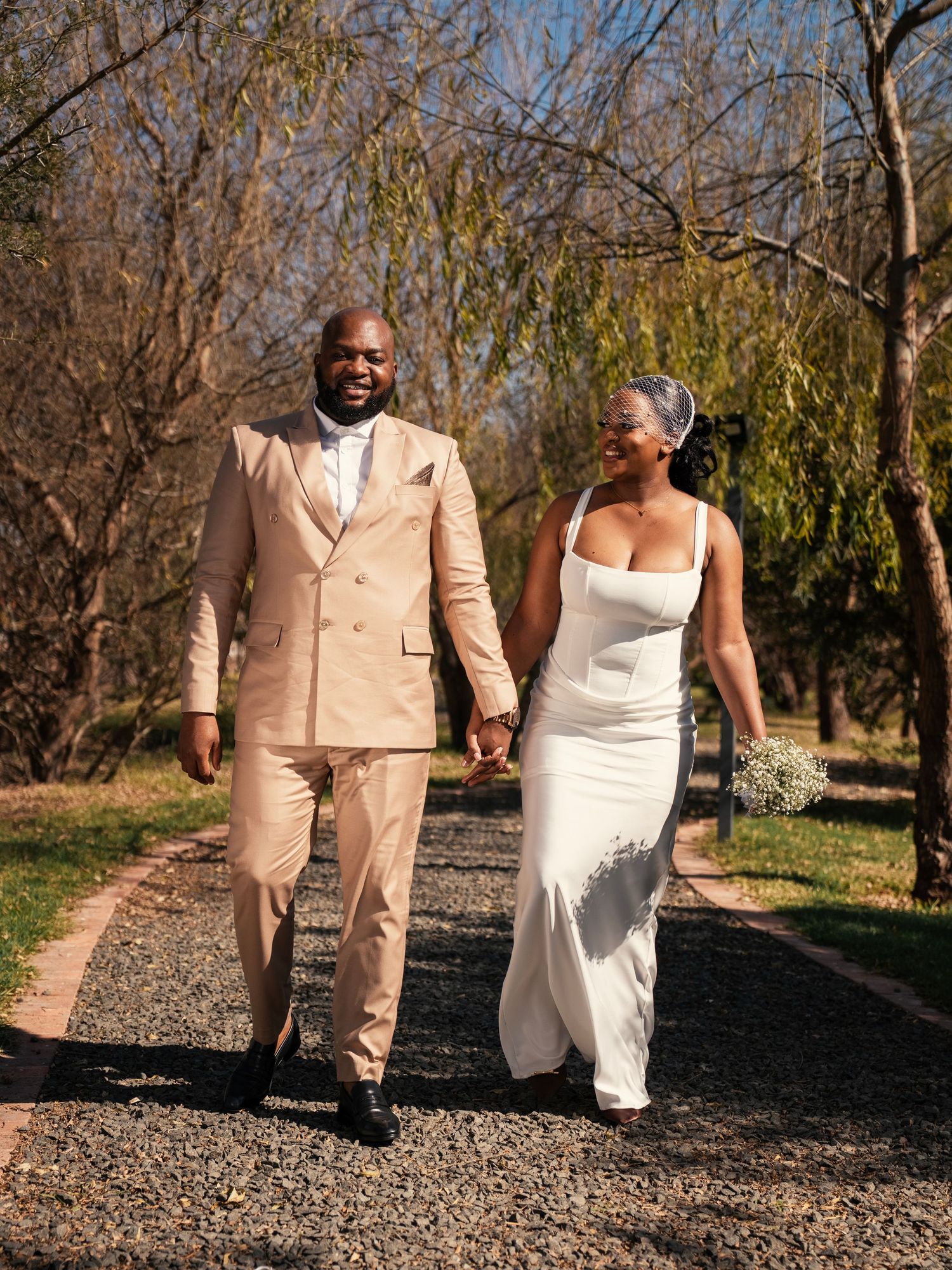Happy couple walks along tree-lined path on wedding day, bride holding baby's breath bouquet. Wedding Photography