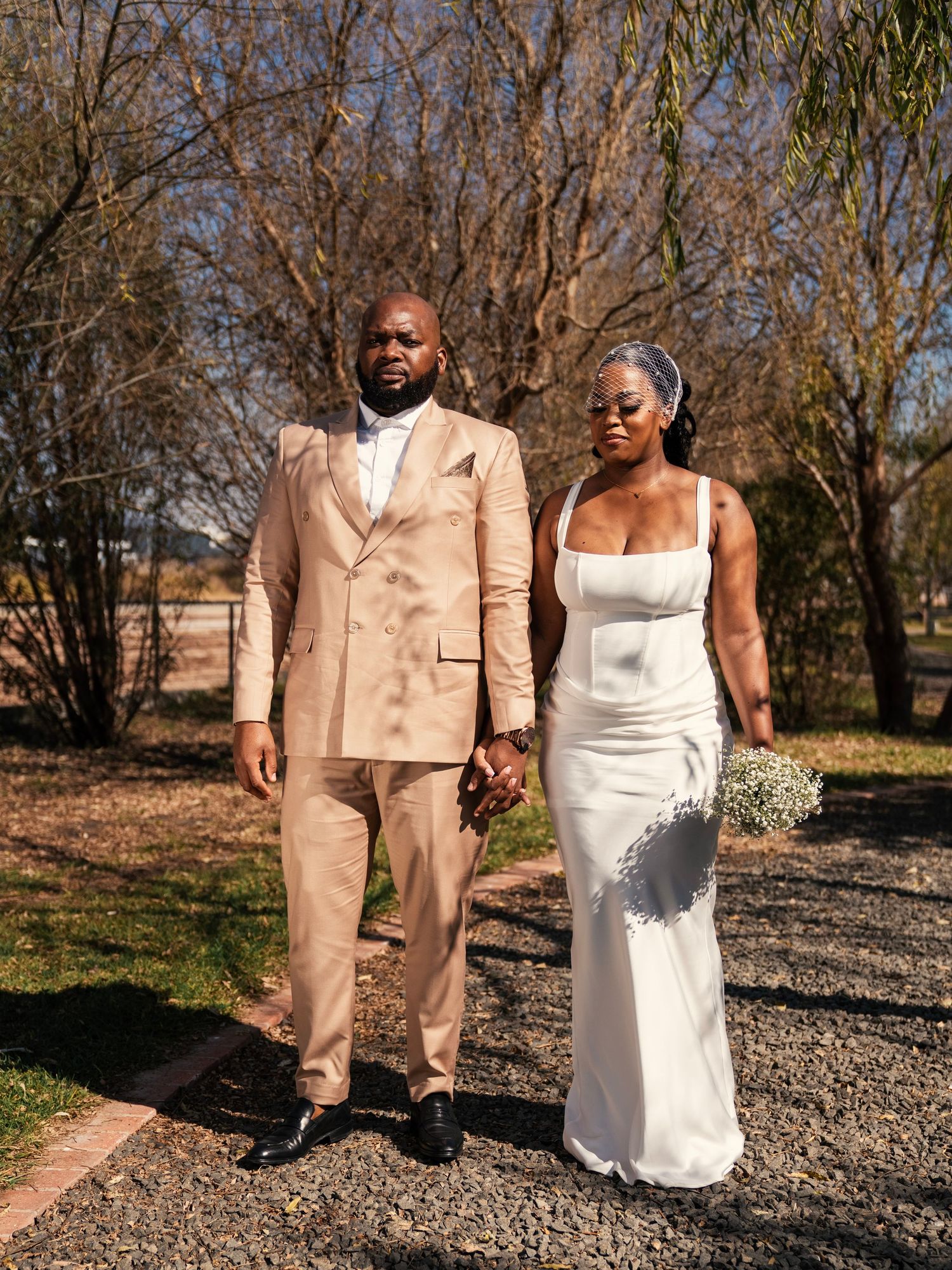 Wedding couple posing hand in hand outdoors, groom in tan double-breasted suit and bride in sleek white dress.
