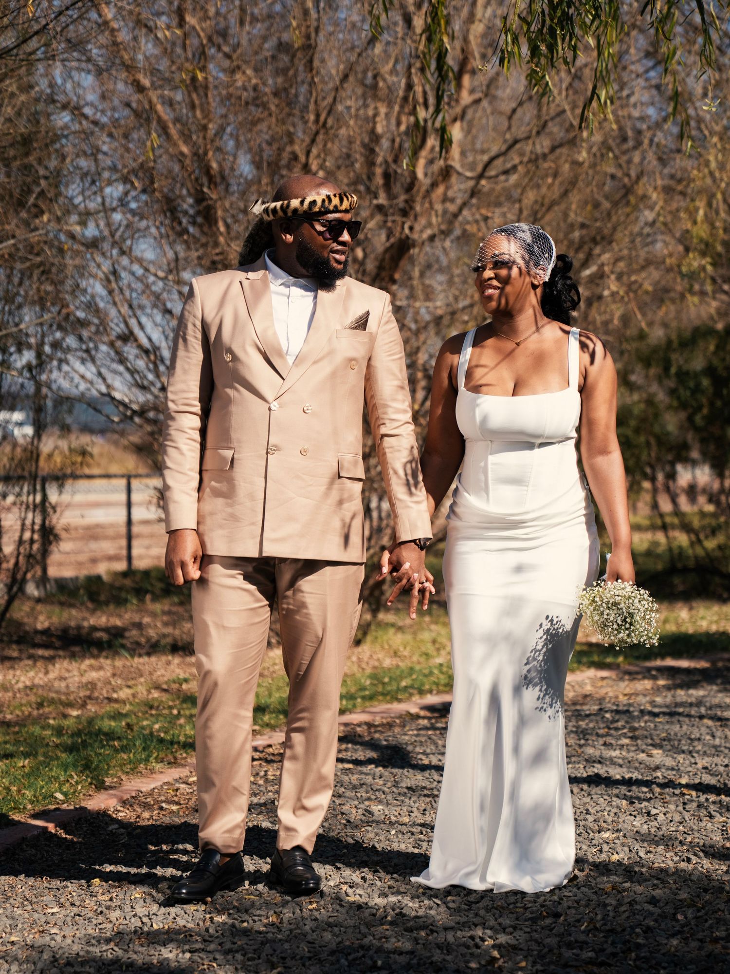 Newlyweds walk hand in hand outdoors, groom in beige suit, bride in white gown. Wedding Photography