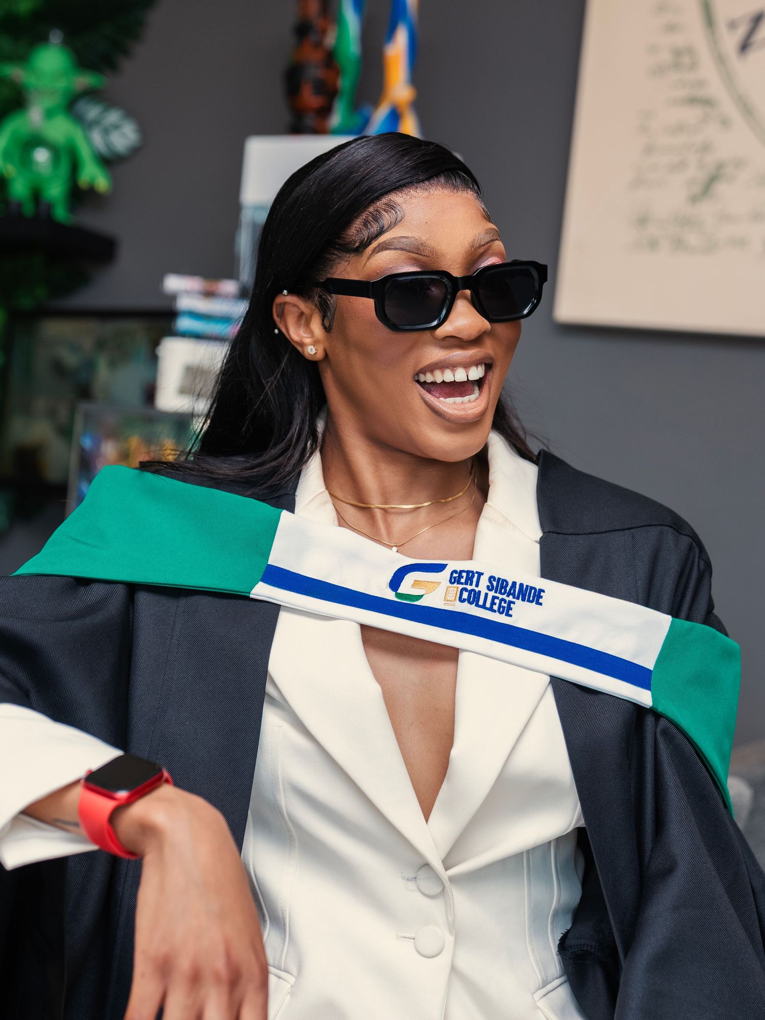 Smiling graduate in black gown with Gert Sibande College hood wearing sunglasses indoors. Babyshower Photoshoot