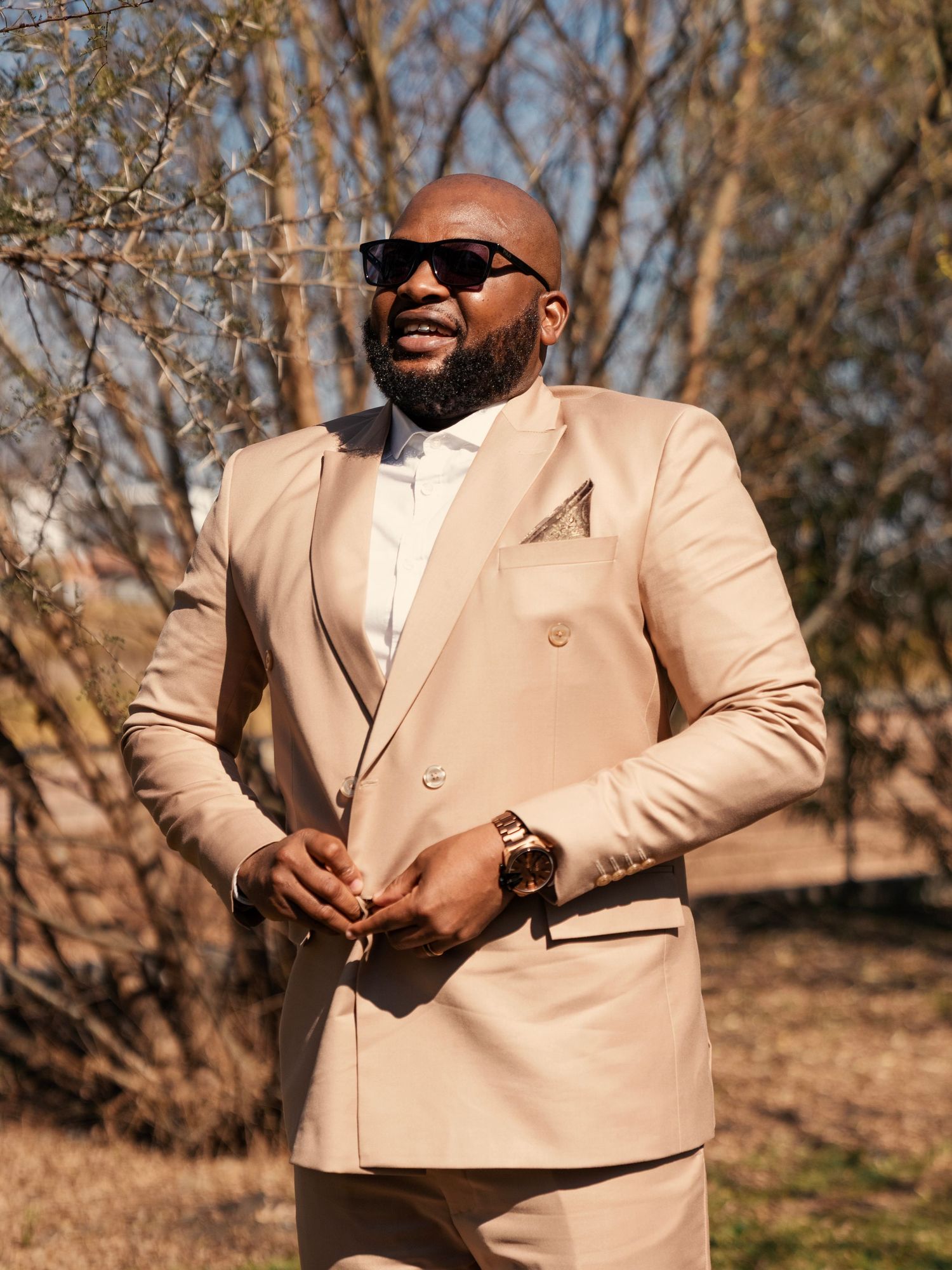A bride in a beige double-breasted suit and sunglasses poses outdoors among bare trees. Wedding Photography