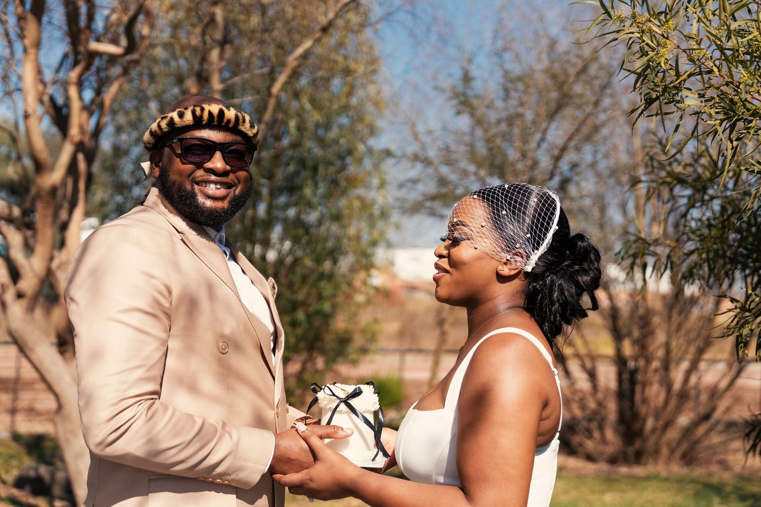 A joyful Black couple holds hands outdoors, the groom in a tan suit and the bride in a white dress holding a bouquet. Wedding Photography
