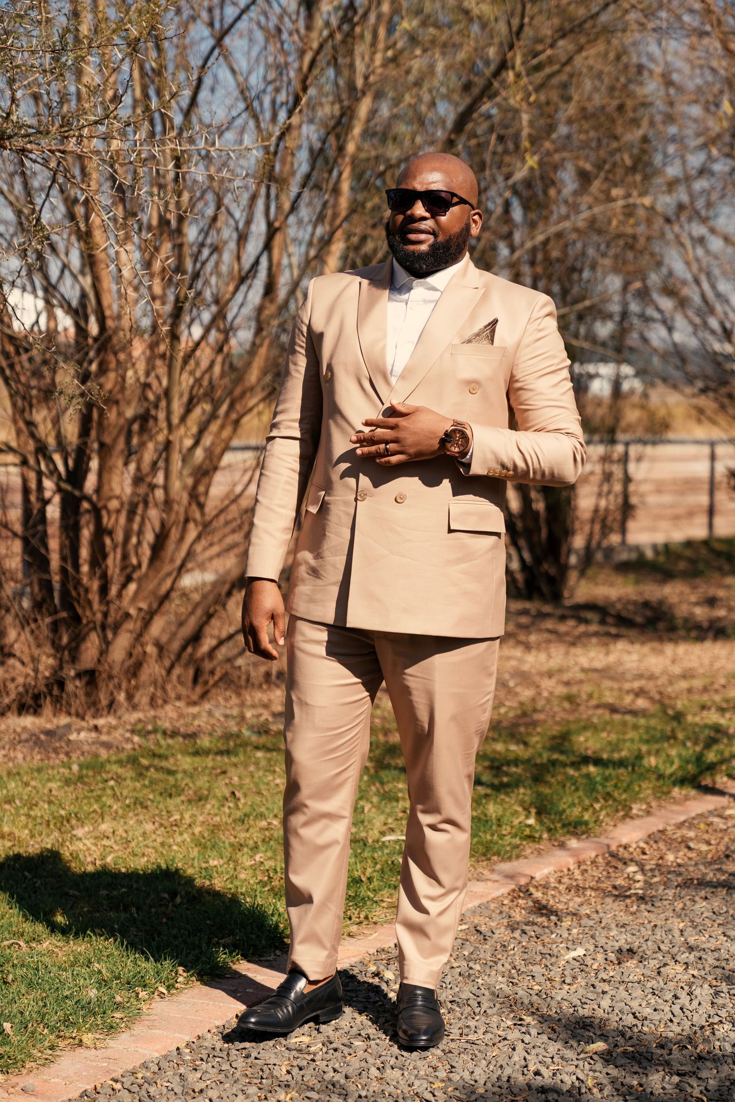 A well-dressed Black man in a beige double-breasted suit and sunglasses poses outdoors on a sunny day. Wedding Photography