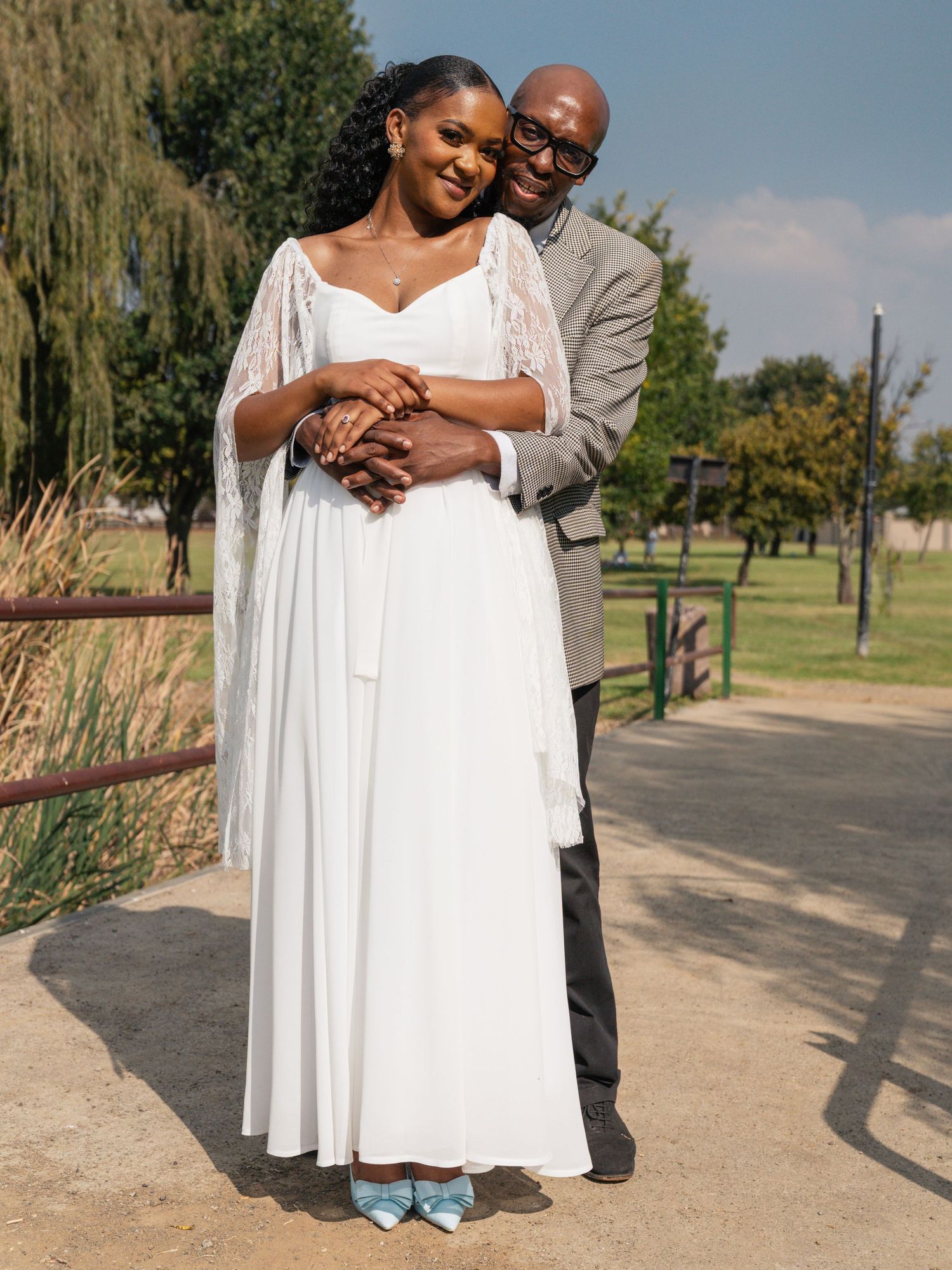 Bride in white gown with lace cape embraced by groom in grey suit outdoors. Wedding Photography