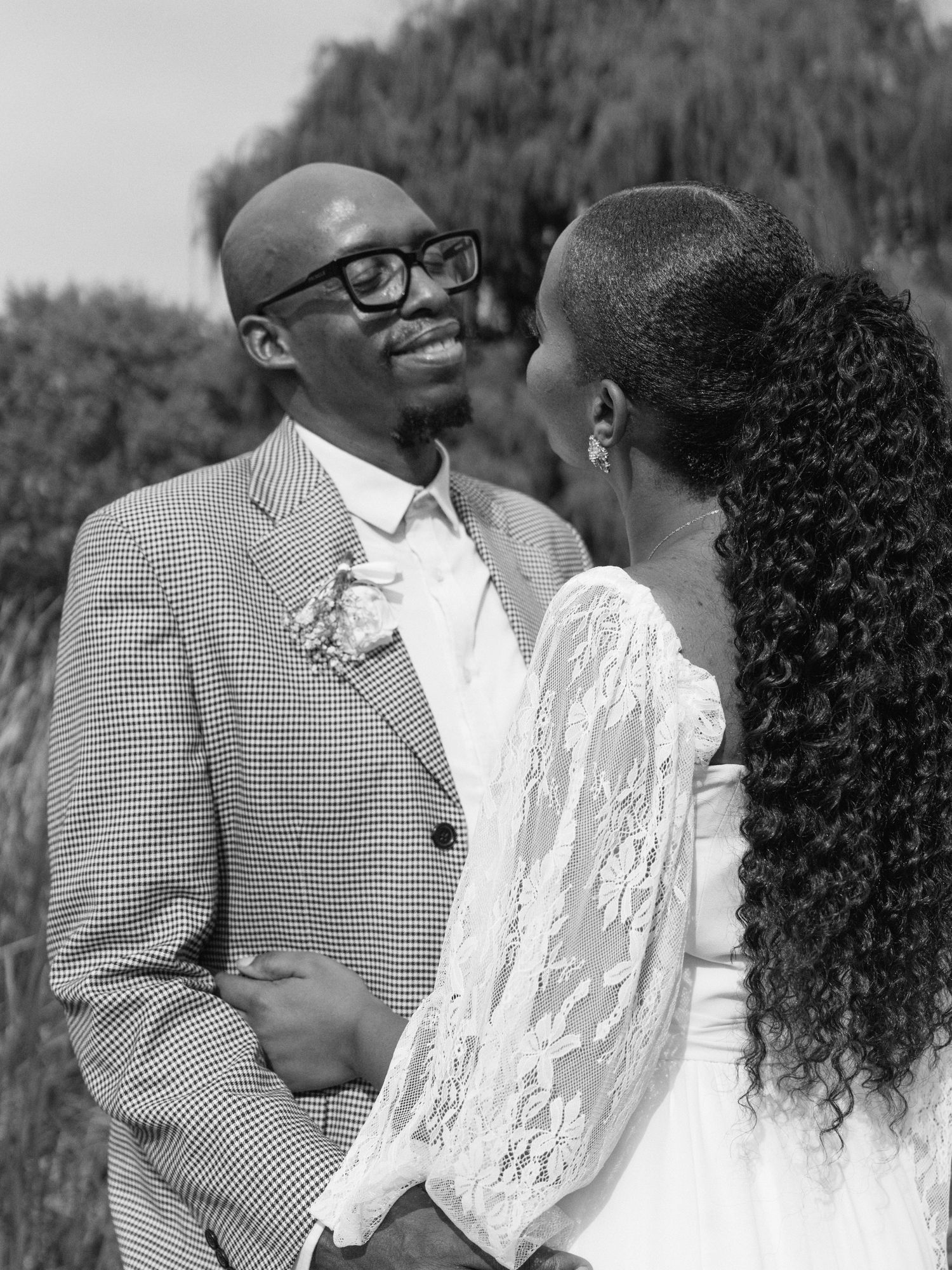 Black and white photo of a smiling groom in a plaid suit gazing at his bride in a lace wedding dress.