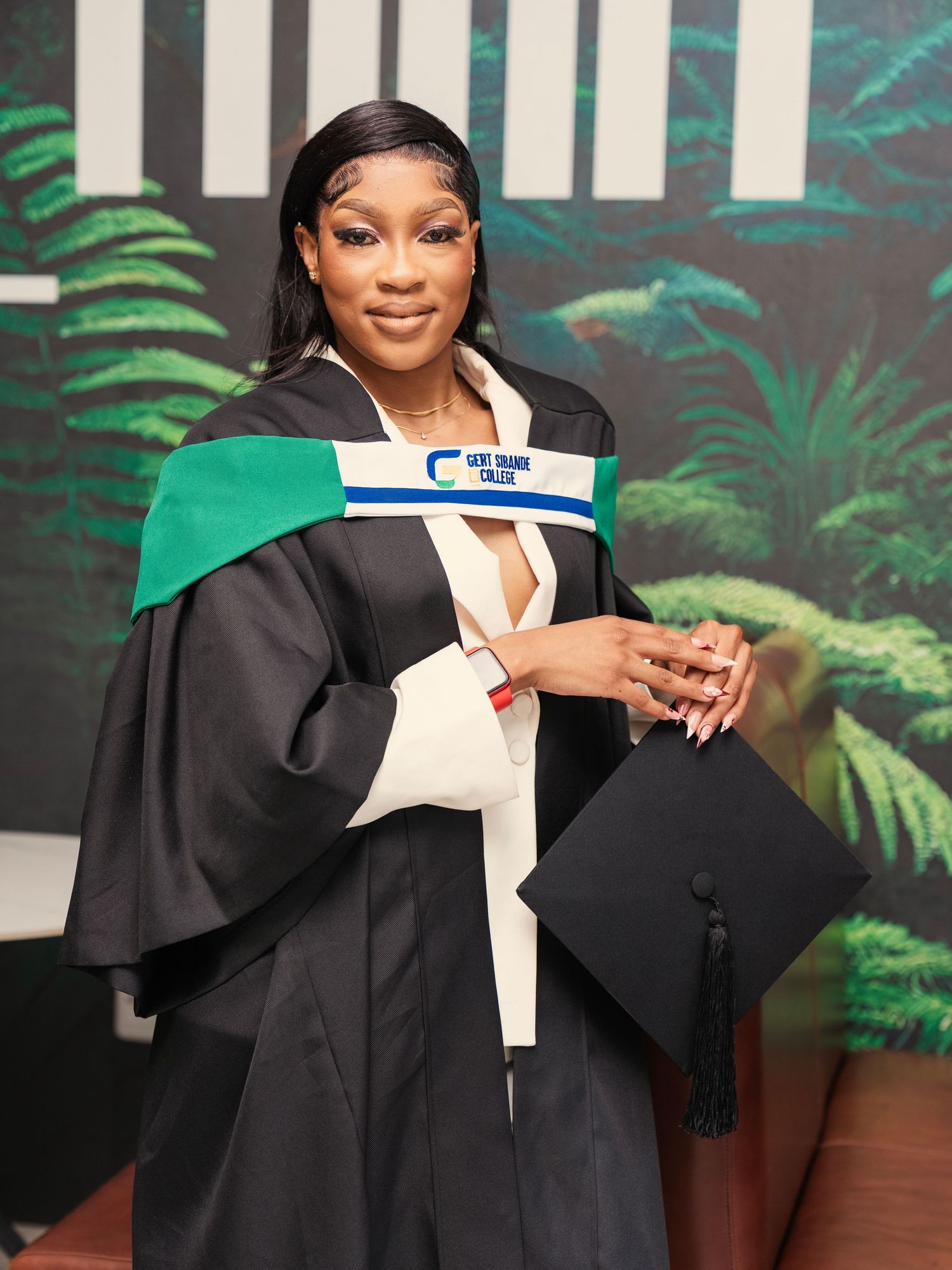 Smiling graduate in black academic gown holding mortarboard cap in front of tropical backdrop. Graduation Shoot Photography