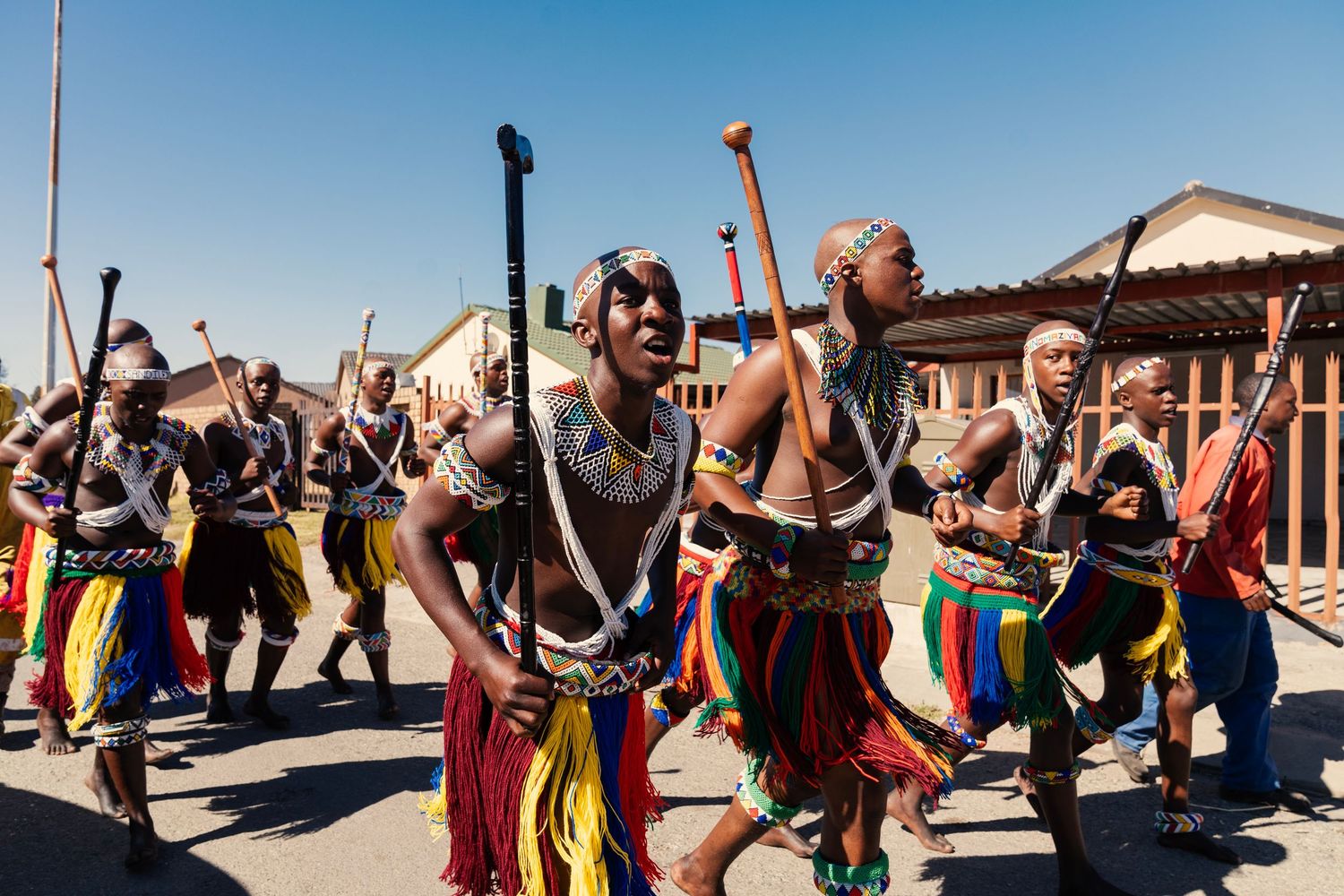 Group of performers in traditional Zulu attire with colorful fringe skirts dancing in a street procession. Event Photography