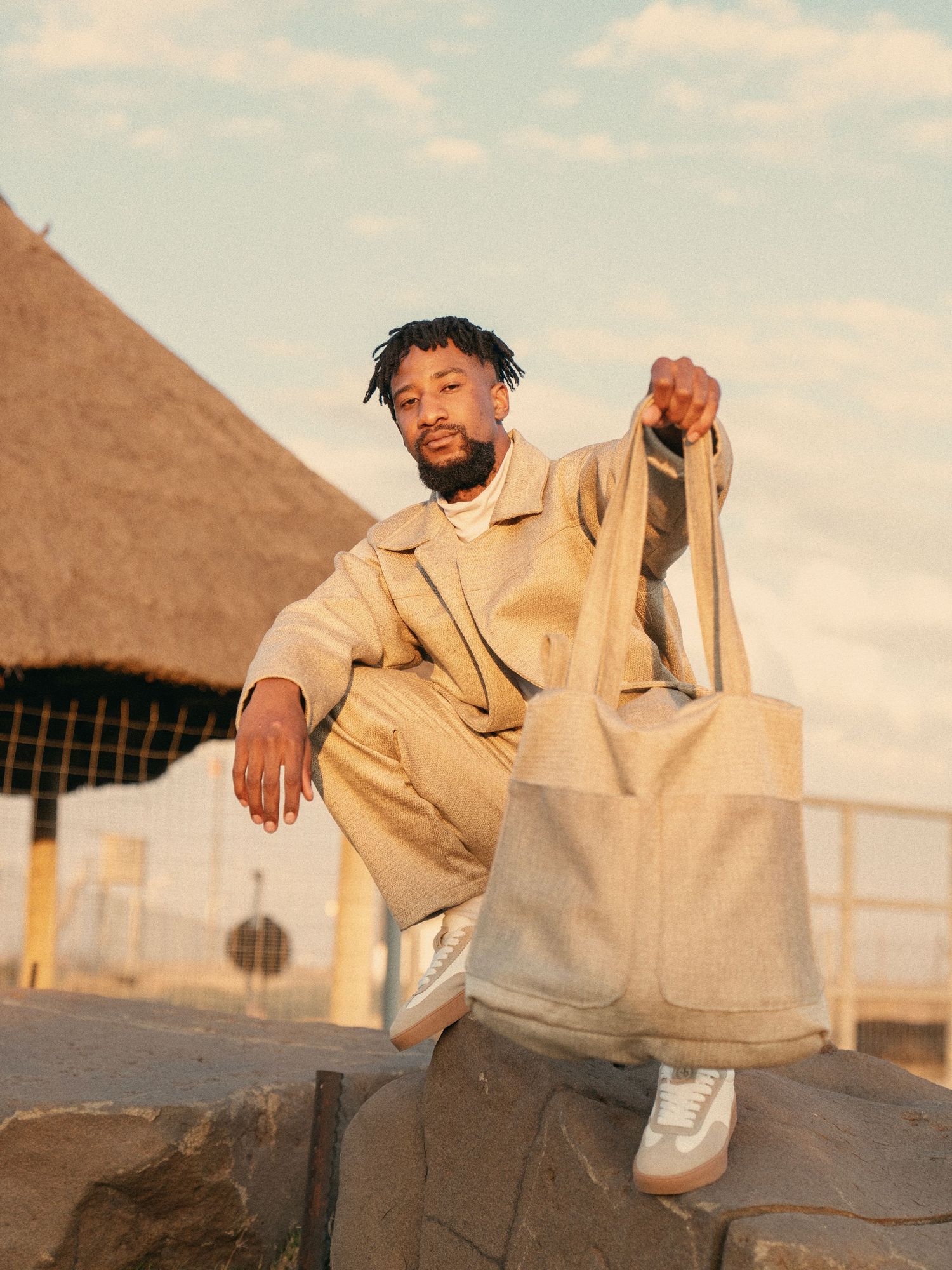 Stylish man in beige outfit holding tote bag sitting on rocks outdoors. Fashion Photography