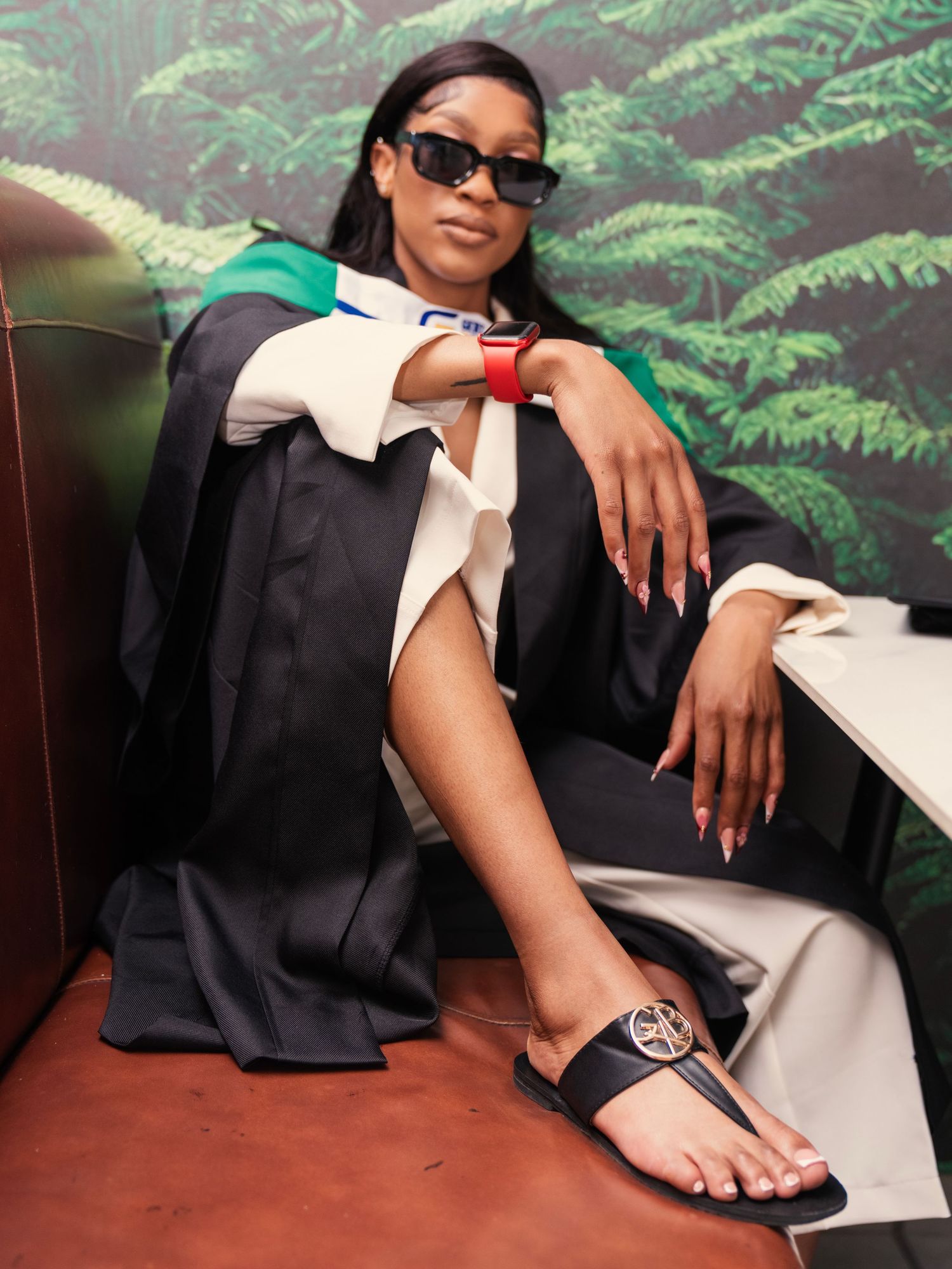 Graduate in black gown and sunglasses seated in booth with tropical green backdrop. Babyshower Photoshoot