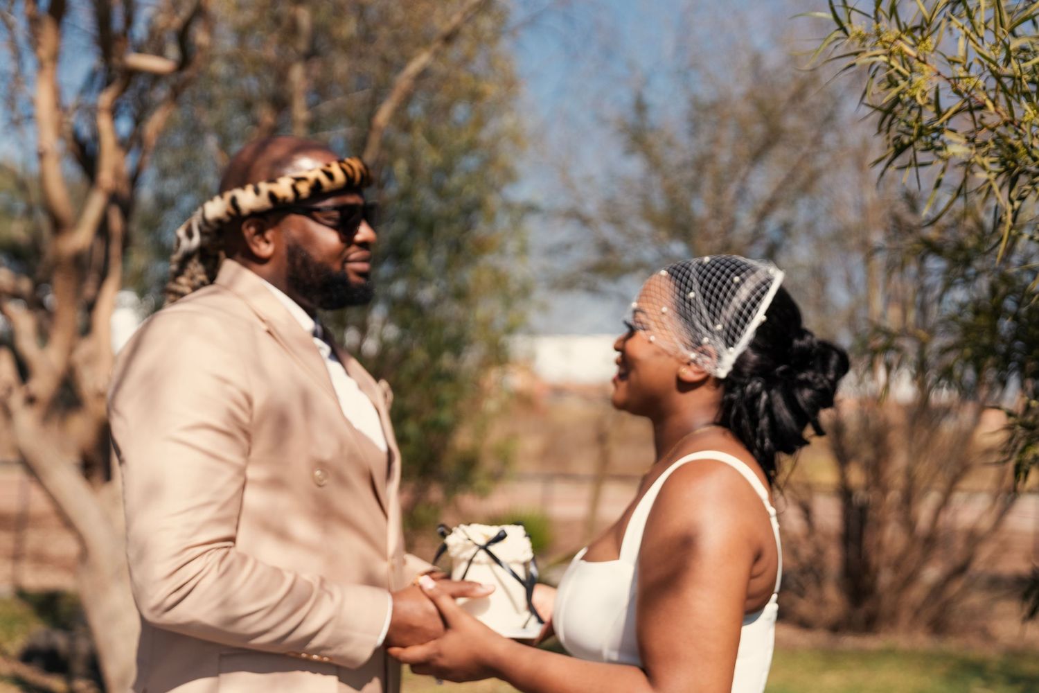 A joyful Black couple holds hands outdoors, the groom in a tan suit and the bride in a white dress holding a bouquet. Wedding Photography