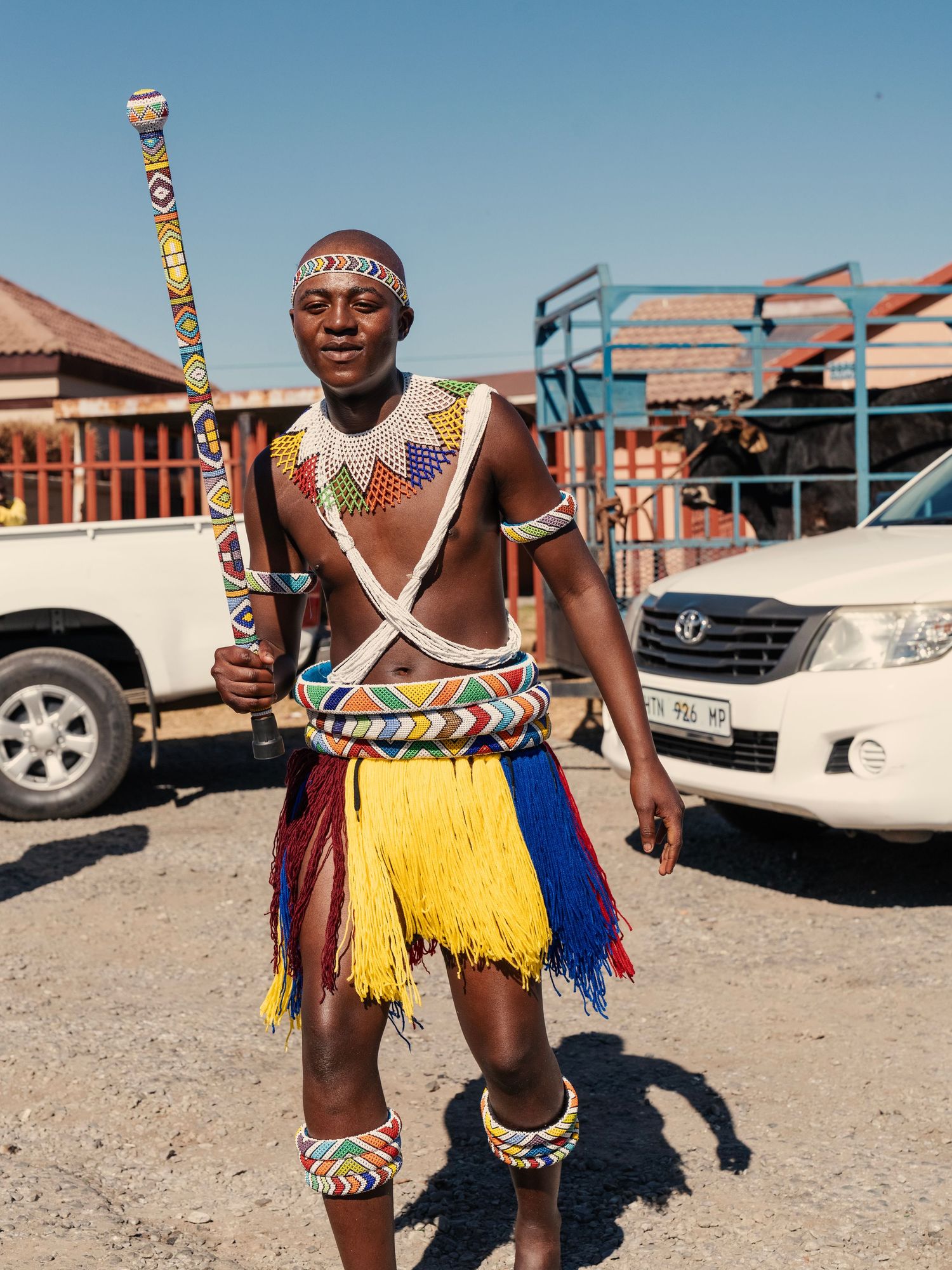 Man in traditional Zulu ceremonial costume with yellow fringe skirt and beaded decorations holding a staff outdoors. Event Photography