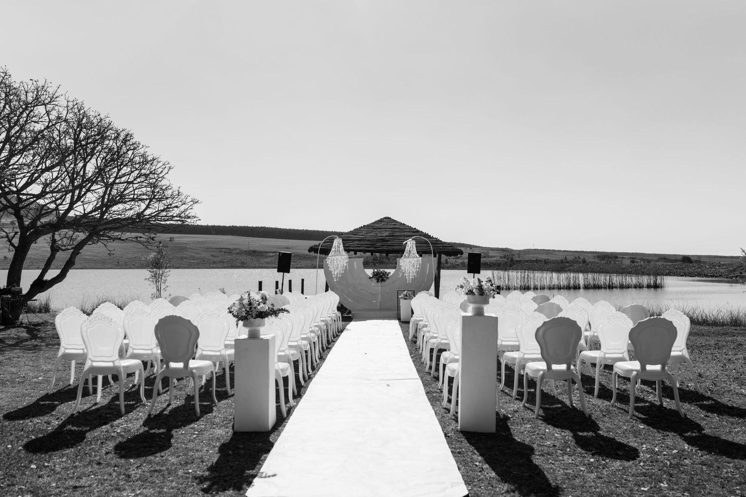 Black and white photo of elegant outdoor wedding ceremony setup with white chairs by water. Wedding Photography