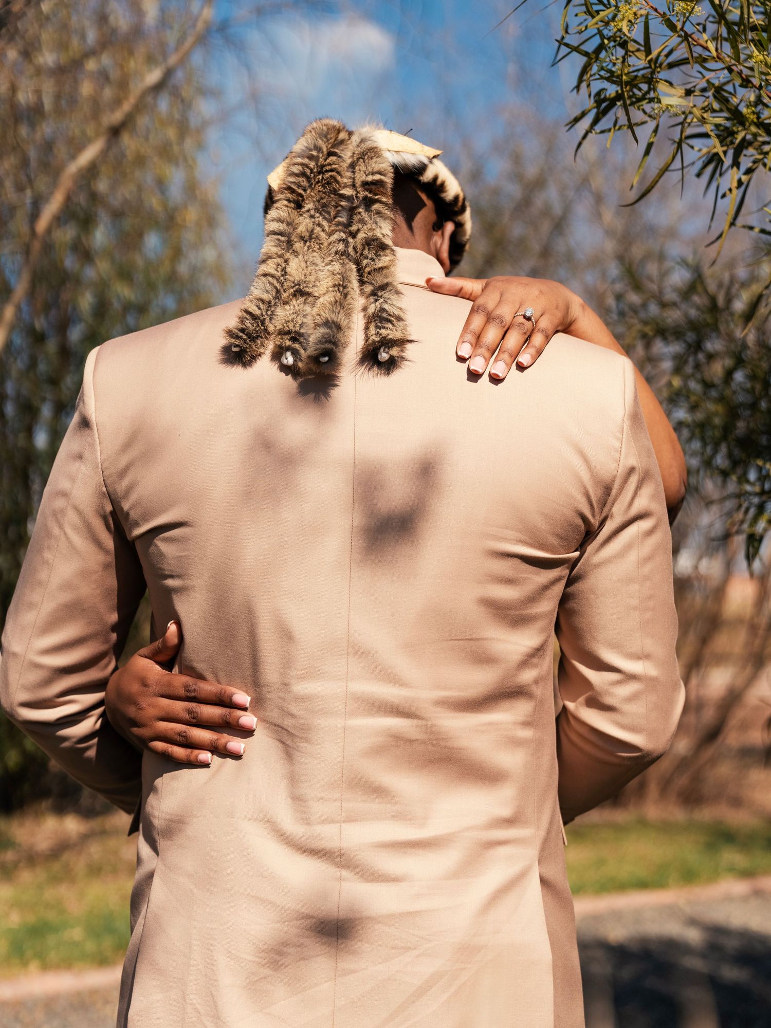 Couple embracing outdoors, man in beige suit with decorative braided hair accessory, woman's hand on his shoulder. Wedding Photography