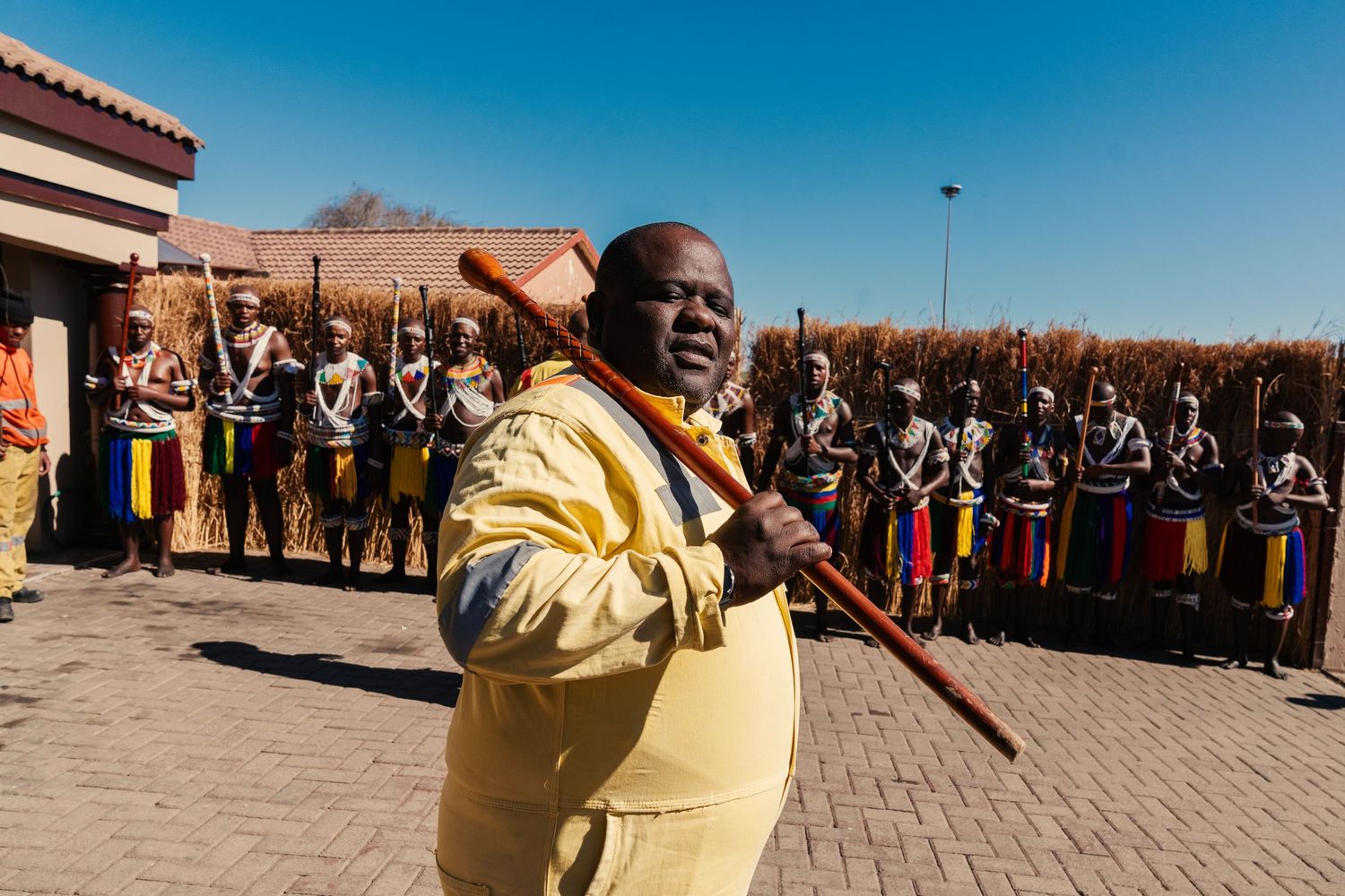 Man in yellow robe leads traditional African ceremony with warriors holding sticks in background. Event Photography