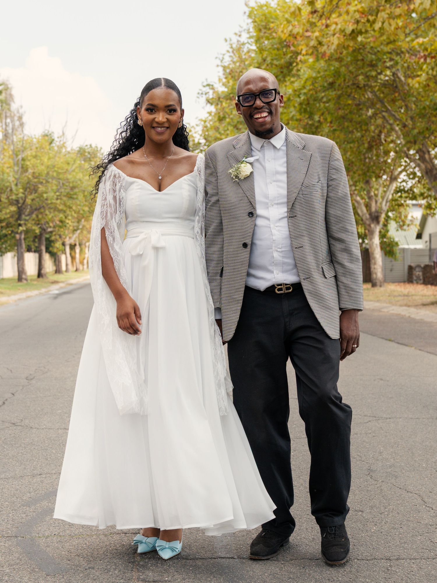Smiling newlywed couple posing on a sunny street, bride in white midi dress with blue heels and groom in grey suit.