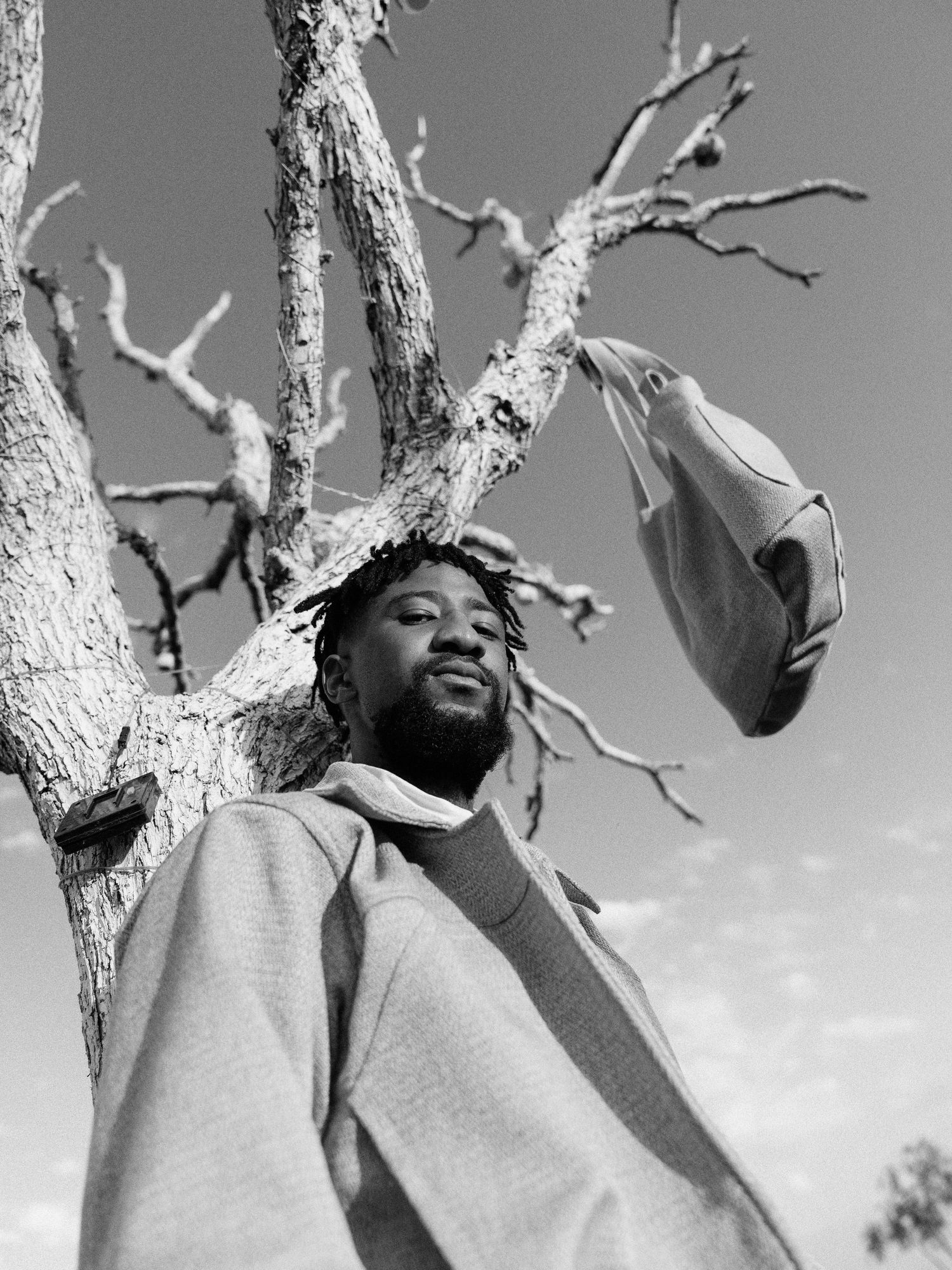 Black and white portrait of a bearded man leaning against a bare tree holding a bag overhead. Fashion Photography
