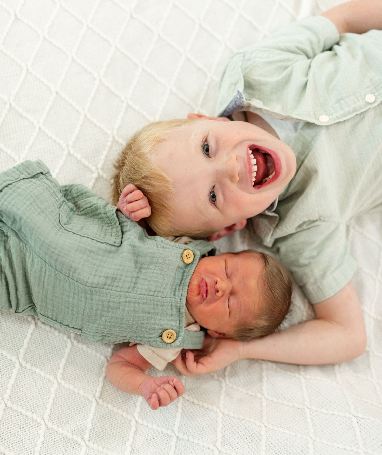 Smiling toddler boy in green shirt lying next to newborn baby sibling on white textured blanket. Newborn Photography Near me