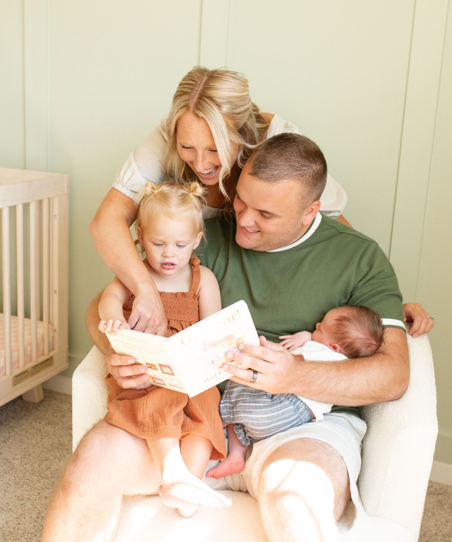 Family of four reading a book together in a nursery, parents smiling at toddler. Ofallon Newborn Photographer