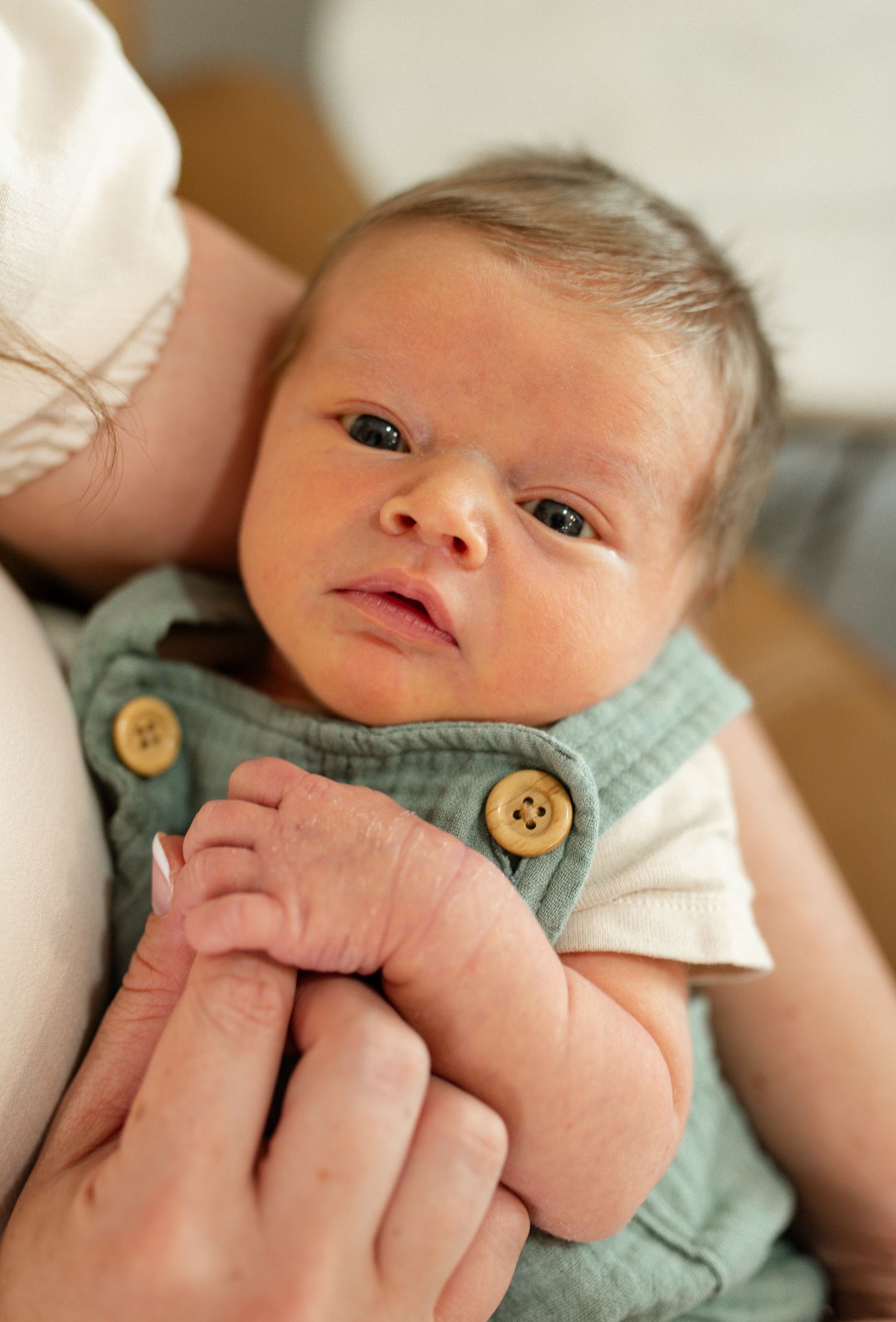 Newborn baby in green overalls being cradled in adult arms, looking alert with dark hair and bright eyes. St Louis Newborn Photography