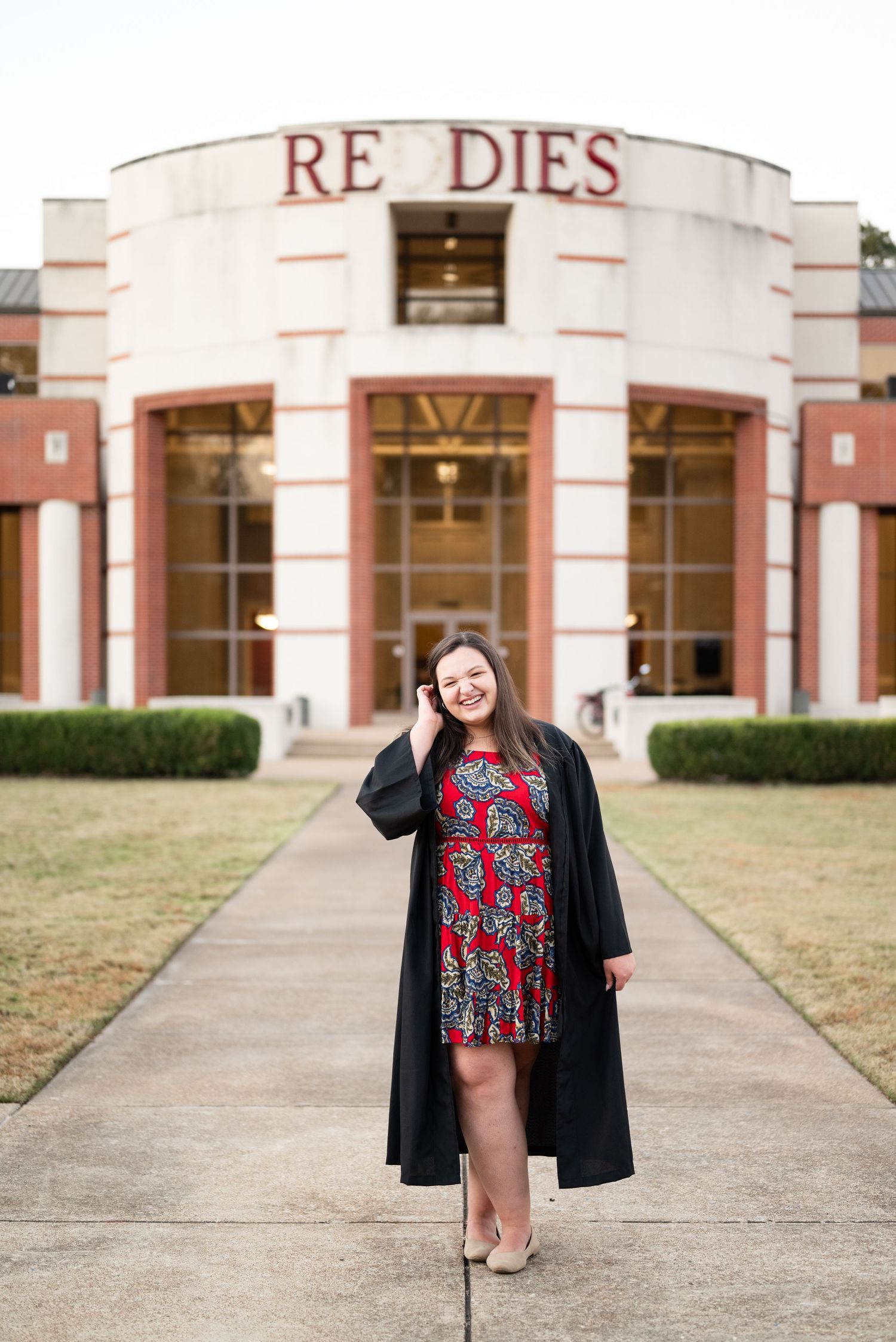 College graduate in a red floral dress and black gown poses in front of a brick building with white columns.