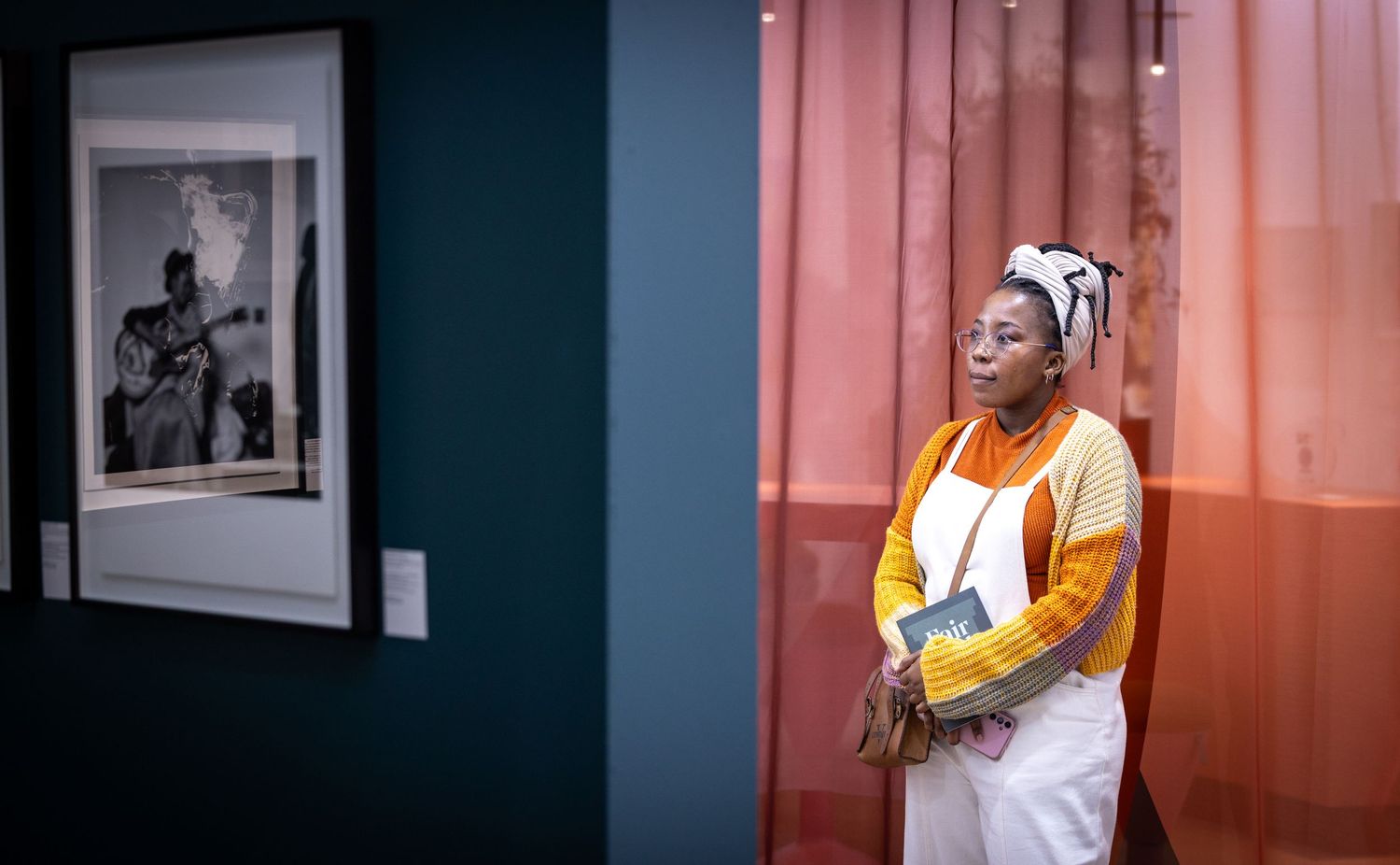 Person wearing a yellow jacket, white pants and head wrap stands against a pink and teal wall with framed artwork.