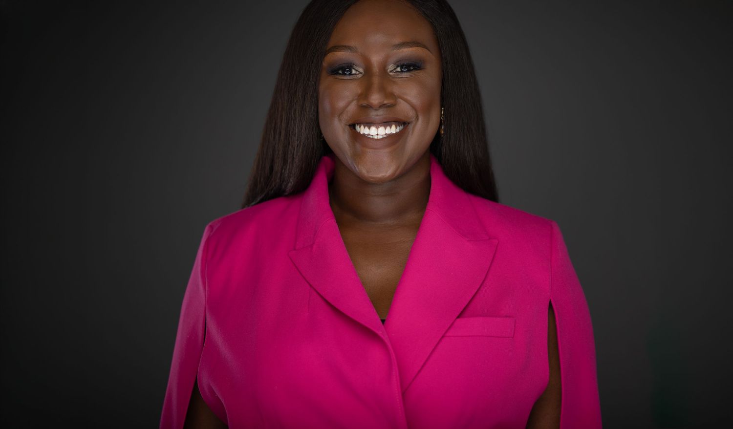 Professional headshot portrait against dark background featuring bright pink blazer and warm smile.