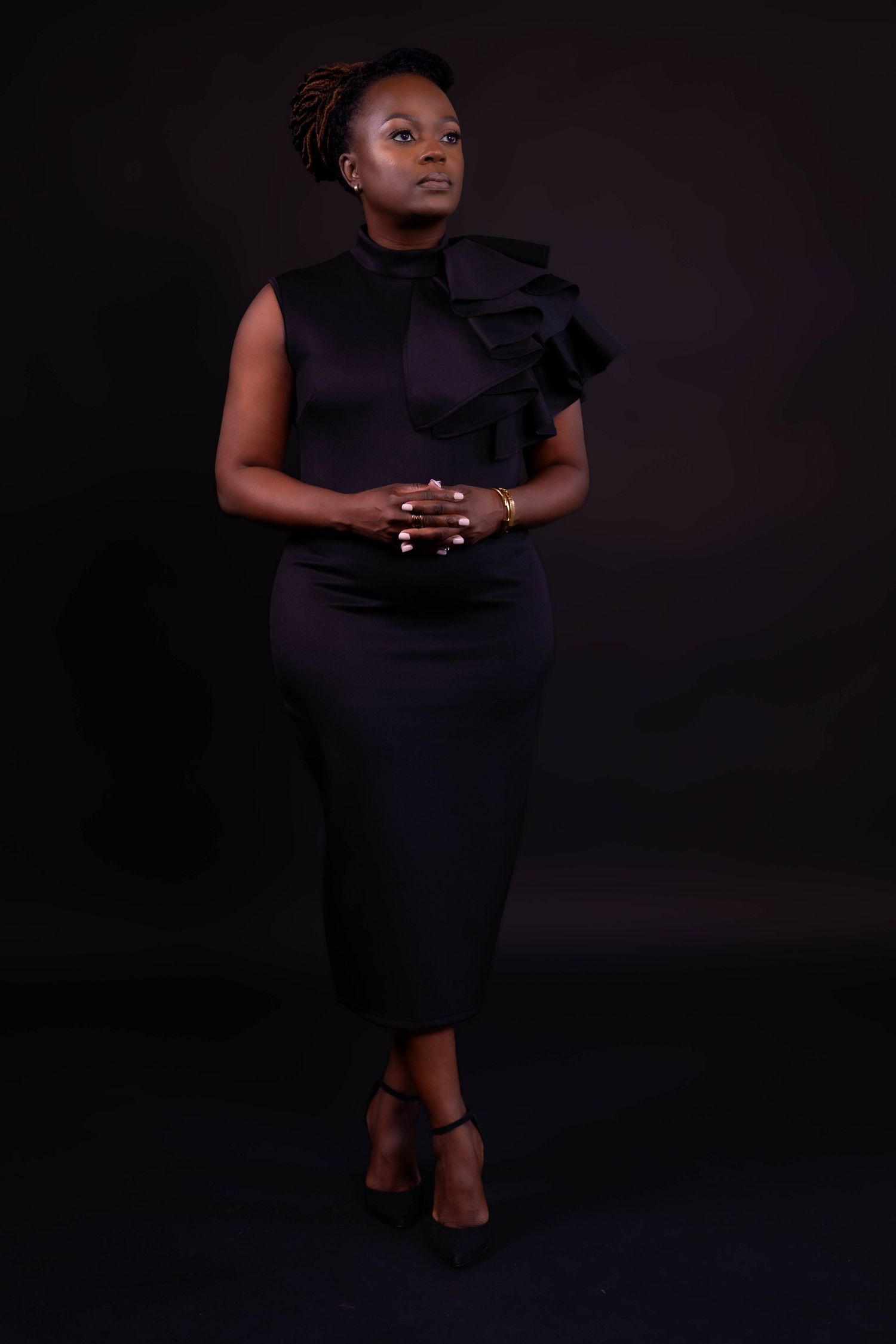 Elegant figure in black formal dress with ruffled shoulder detail poses against dark background in dramatic studio portrait.