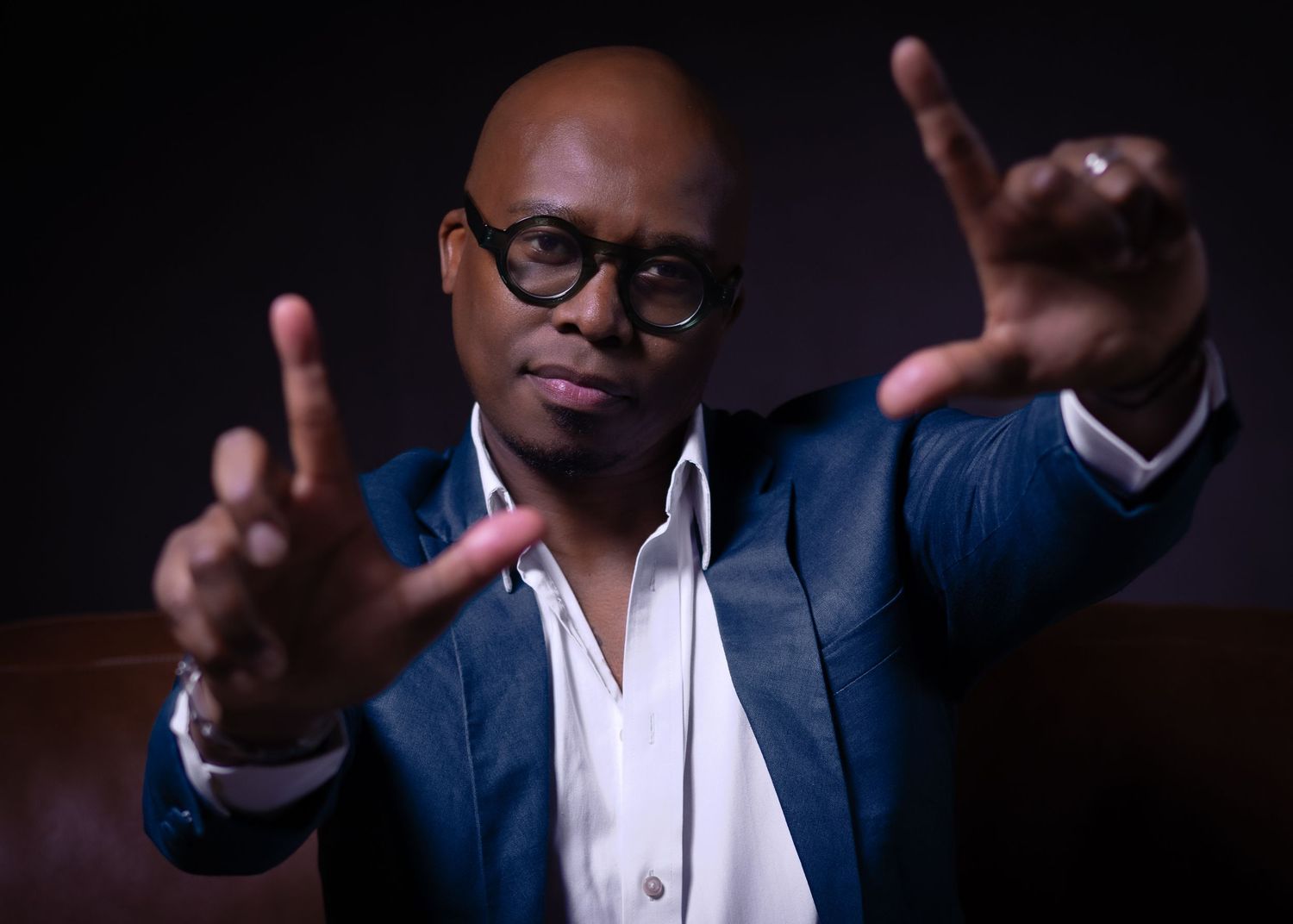 Stylish bald man wearing glasses and a blue blazer gesturing with both hands against a dark background.