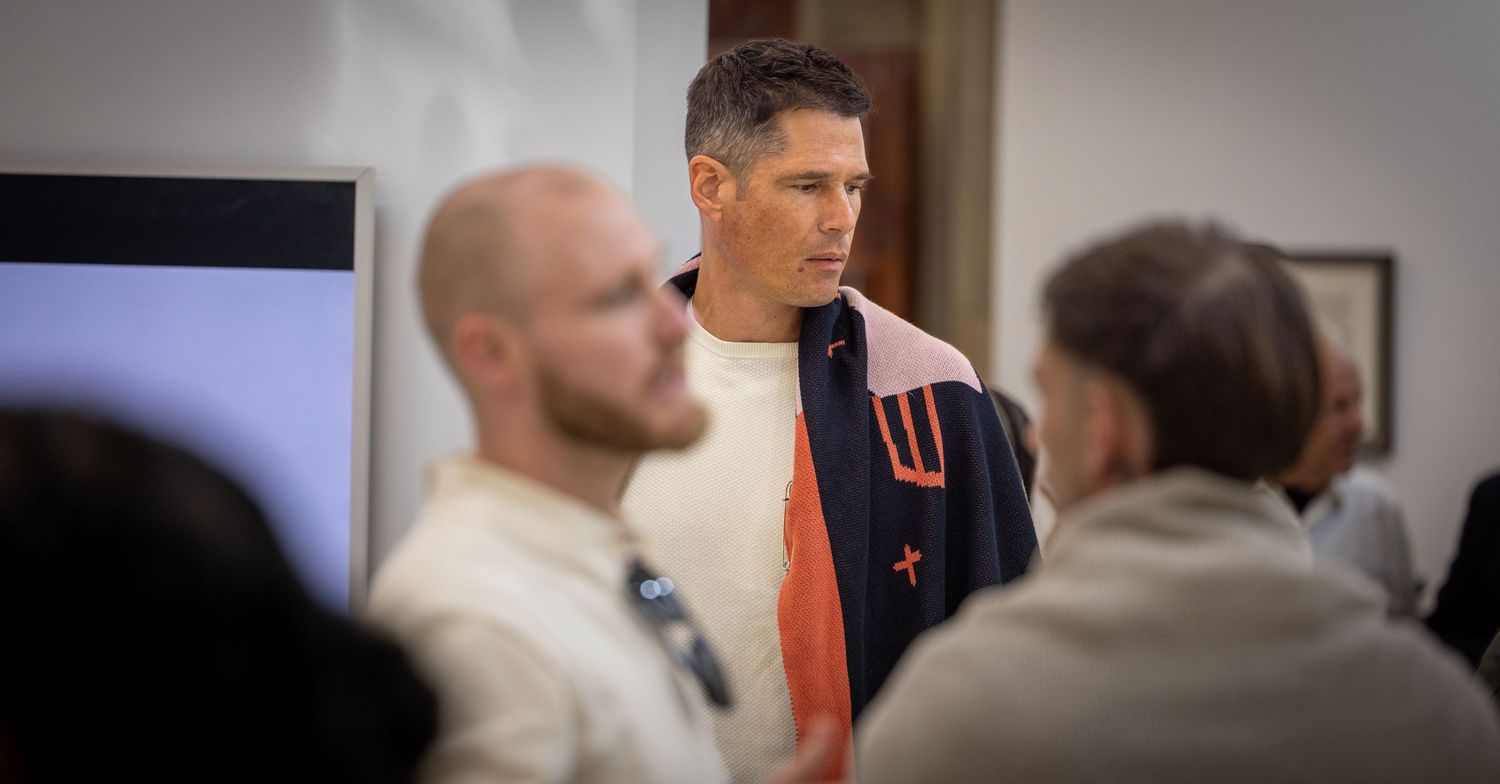 A person in an orange and black varsity jacket speaks with others during what appears to be a meeting or gathering.