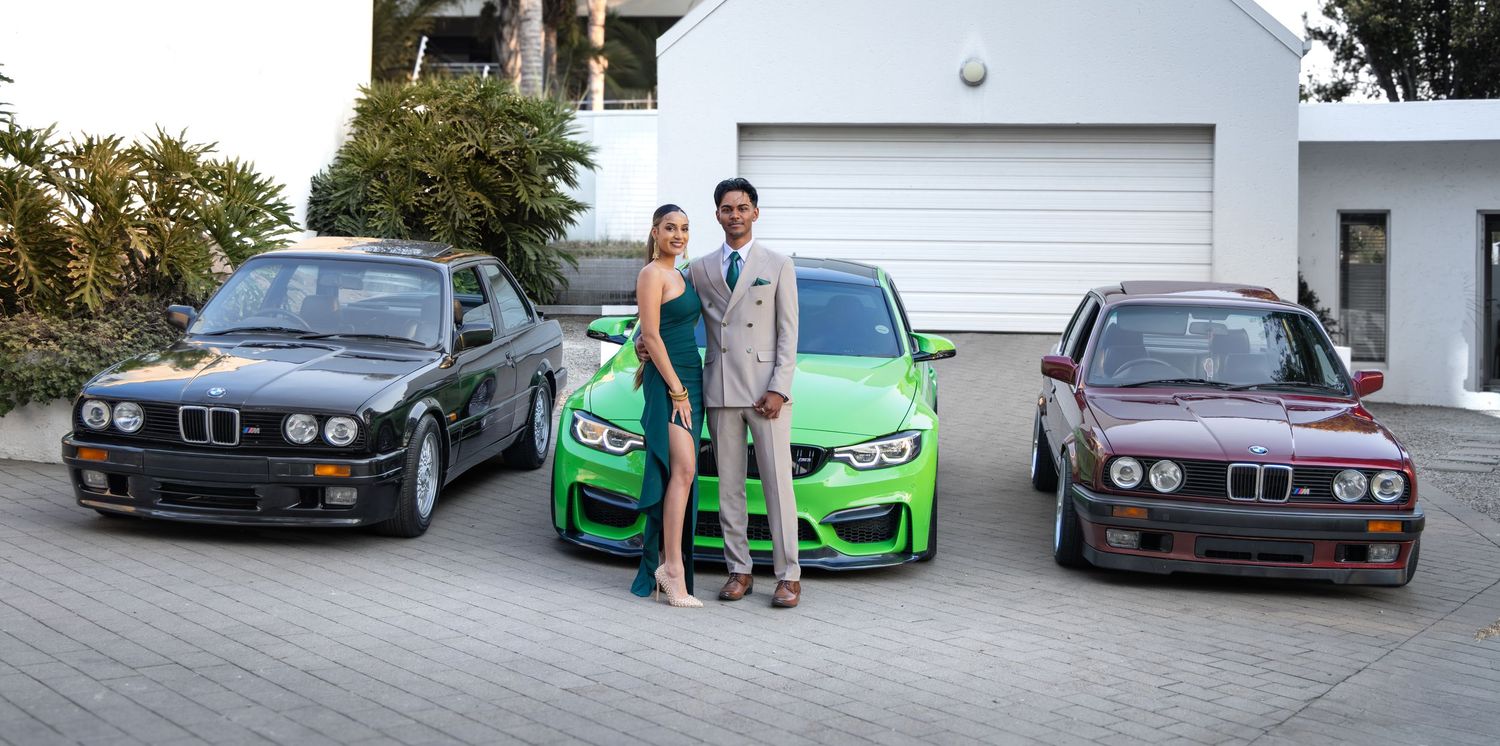 A couple in formal attire poses between three BMW cars, including a bright green M3, on a driveway.