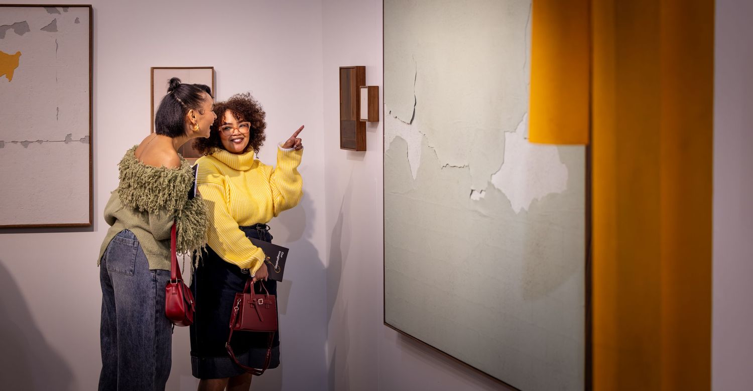 Four people stand close together in casual poses along a white wall with amber lighting.