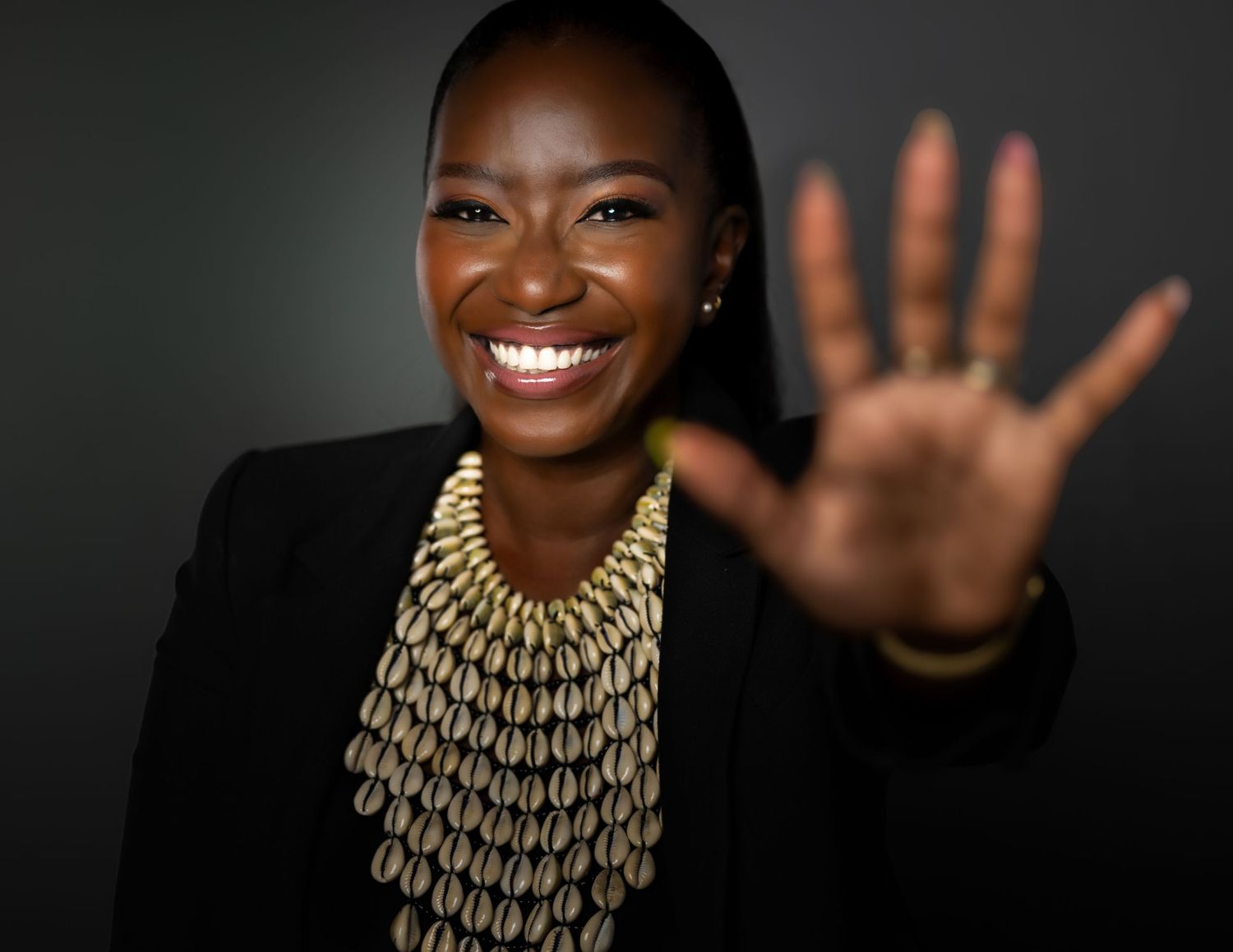 Person wearing black blazer and gold statement necklace raises hand in friendly wave against dark background.