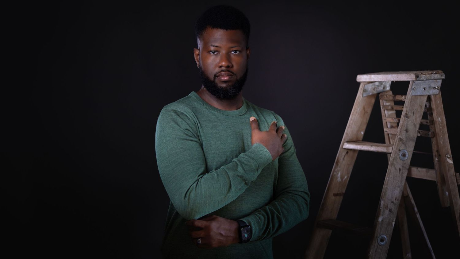 Man in green sweatshirt poses with arm across chest against dark background with wooden ladder visible.