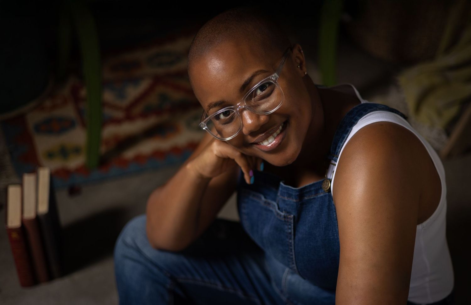Portrait of someone wearing glasses and denim overalls leaning forward with a warm smile in indoor lighting.