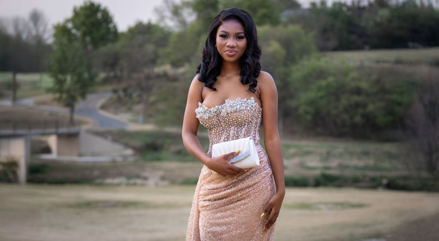 A woman in a peach-colored dress poses gracefully outdoors against a natural landscape background.