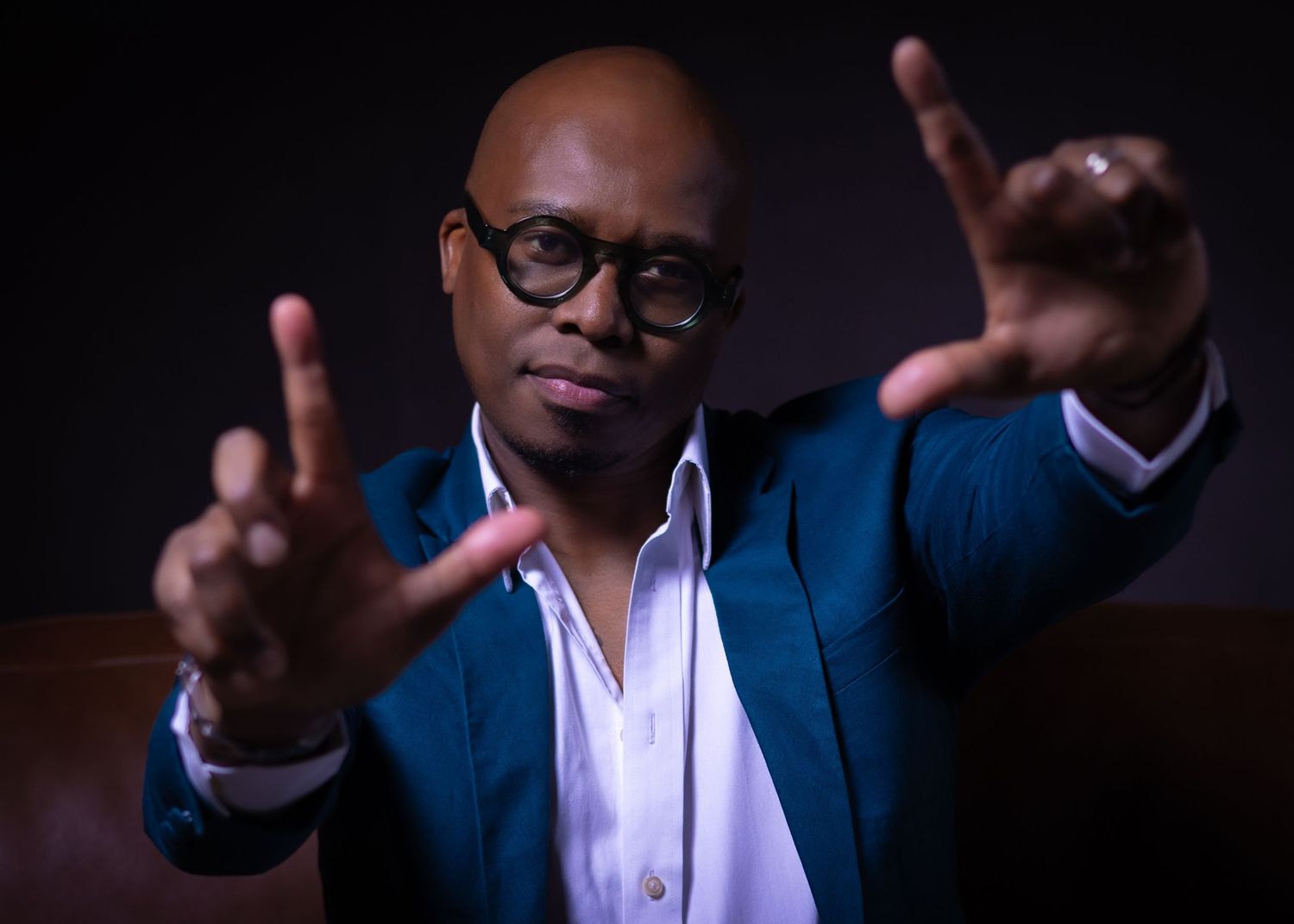 Person in a navy blue suit and white shirt gestures with both hands against a dark background.