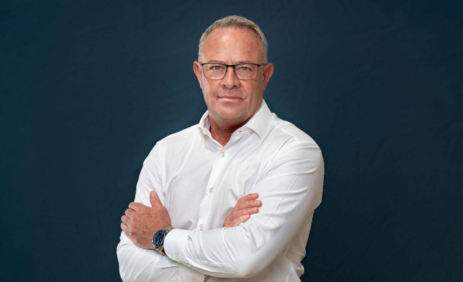 Professional portrait of someone wearing glasses and a white button-down shirt against a dark blue background.